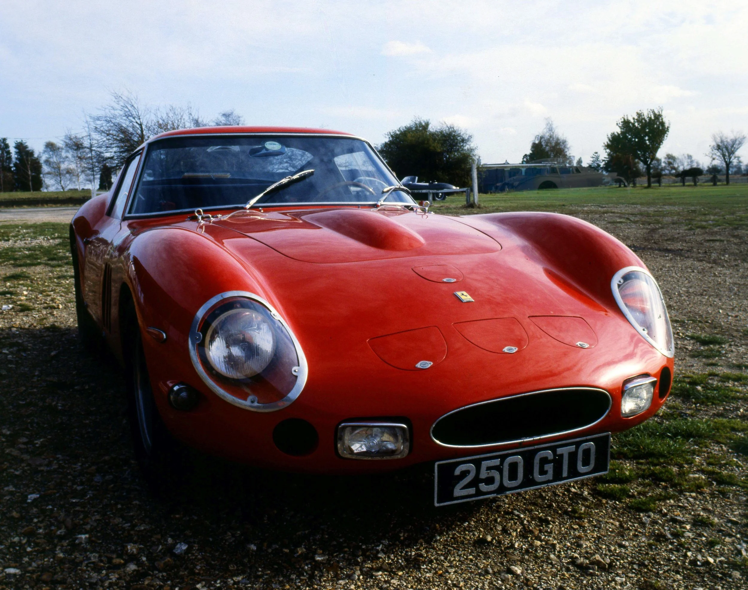 A red vintage Ferrari sports car with a black grille and chrome accents parked outdoors on a gravel surface, with trees and a bus in the background under a partly cloudy sky.