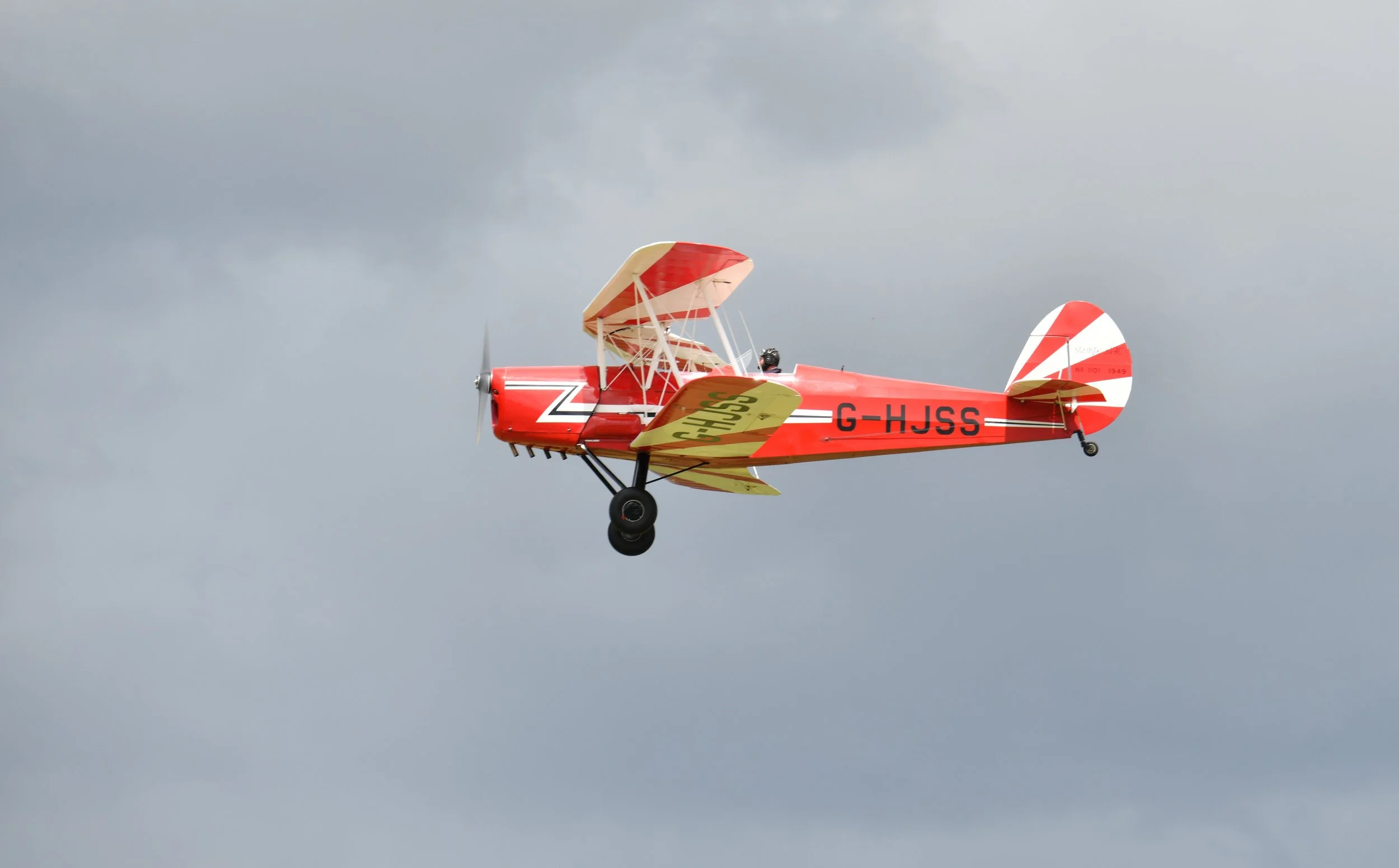 A small red and yellow biplane flying in a cloudy sky.