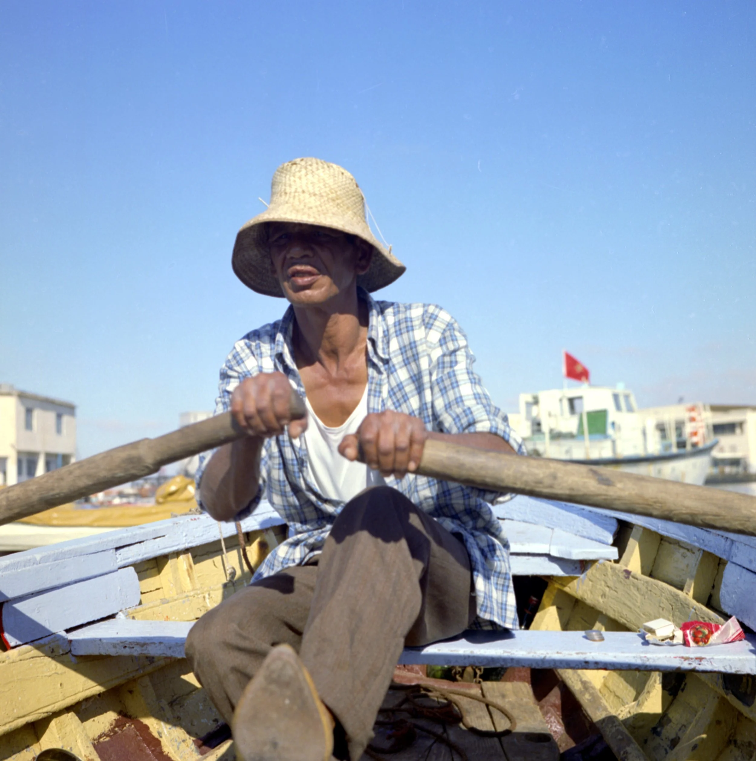 A person wearing a straw hat and plaid shirt rowing a small boat, with a cityscape in the background, under a clear blue sky.