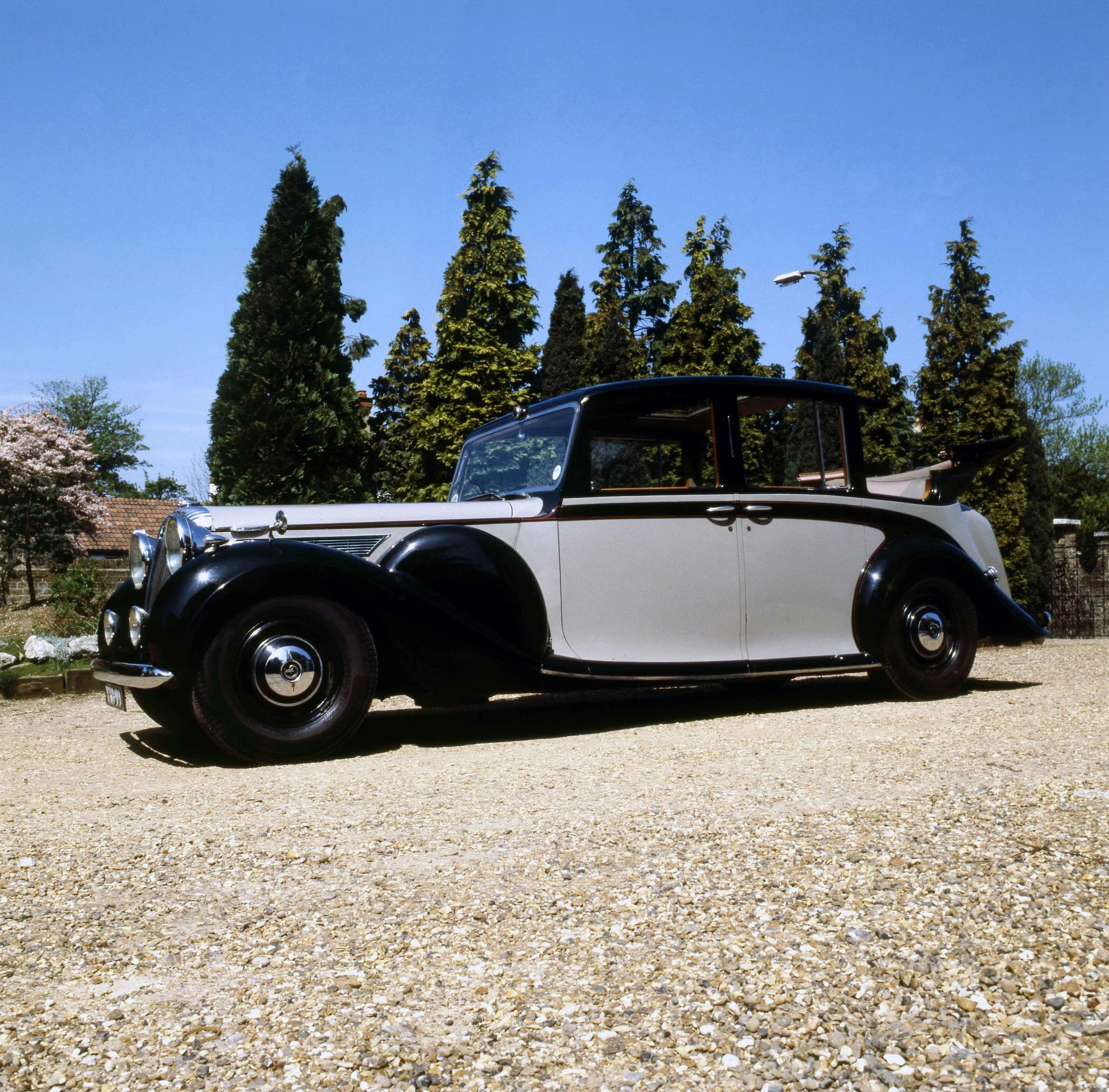 Vintage black and white car parked on a gravel driveway, trees with green foliage in the background, and a bright blue sky.