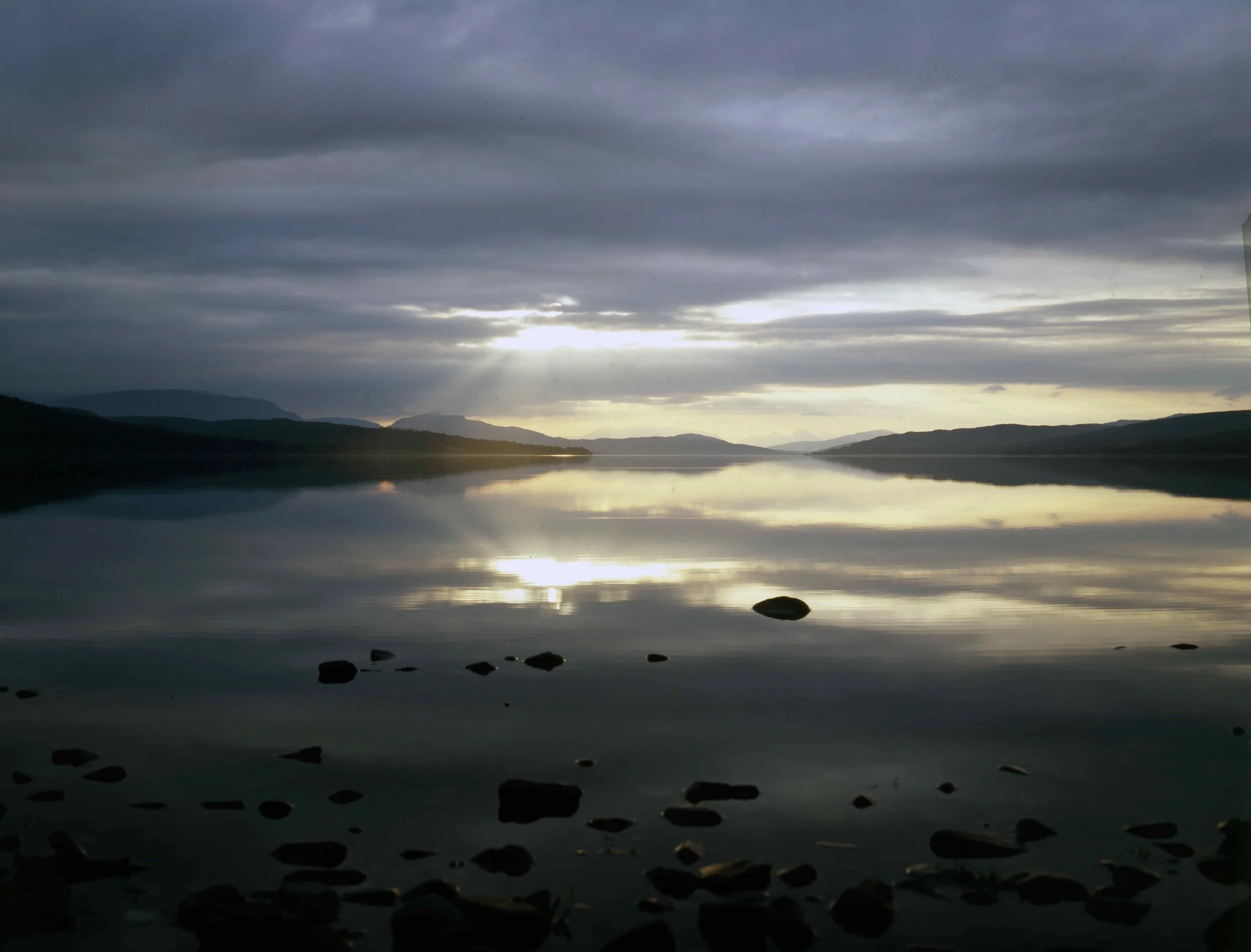 A calm lake with reflections of dark cloudy sky and distant mountains, with rocks scattered along the shoreline.