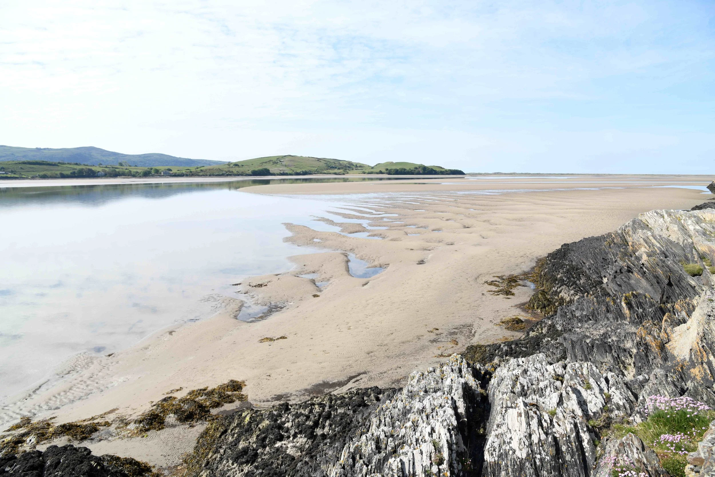 A coastal scene with sandy beach, rocky foreground, calm water reflecting the blue sky, and green hills in the background.