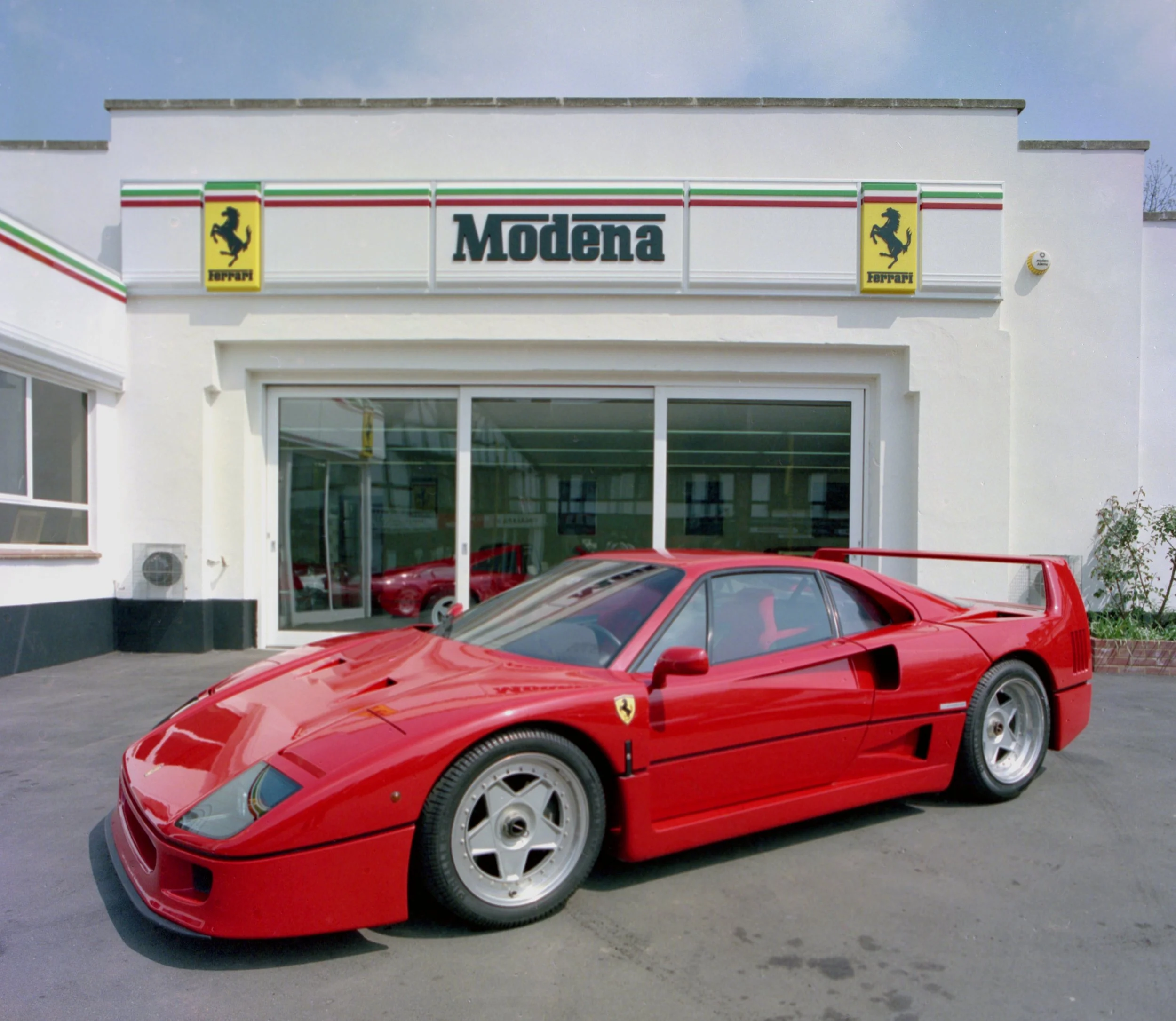 A red Ferrari sports car is parked outside a Ferrari dealership named Modena, which features a white building with Ferrari logos and decorative stripes in green, white, and red.