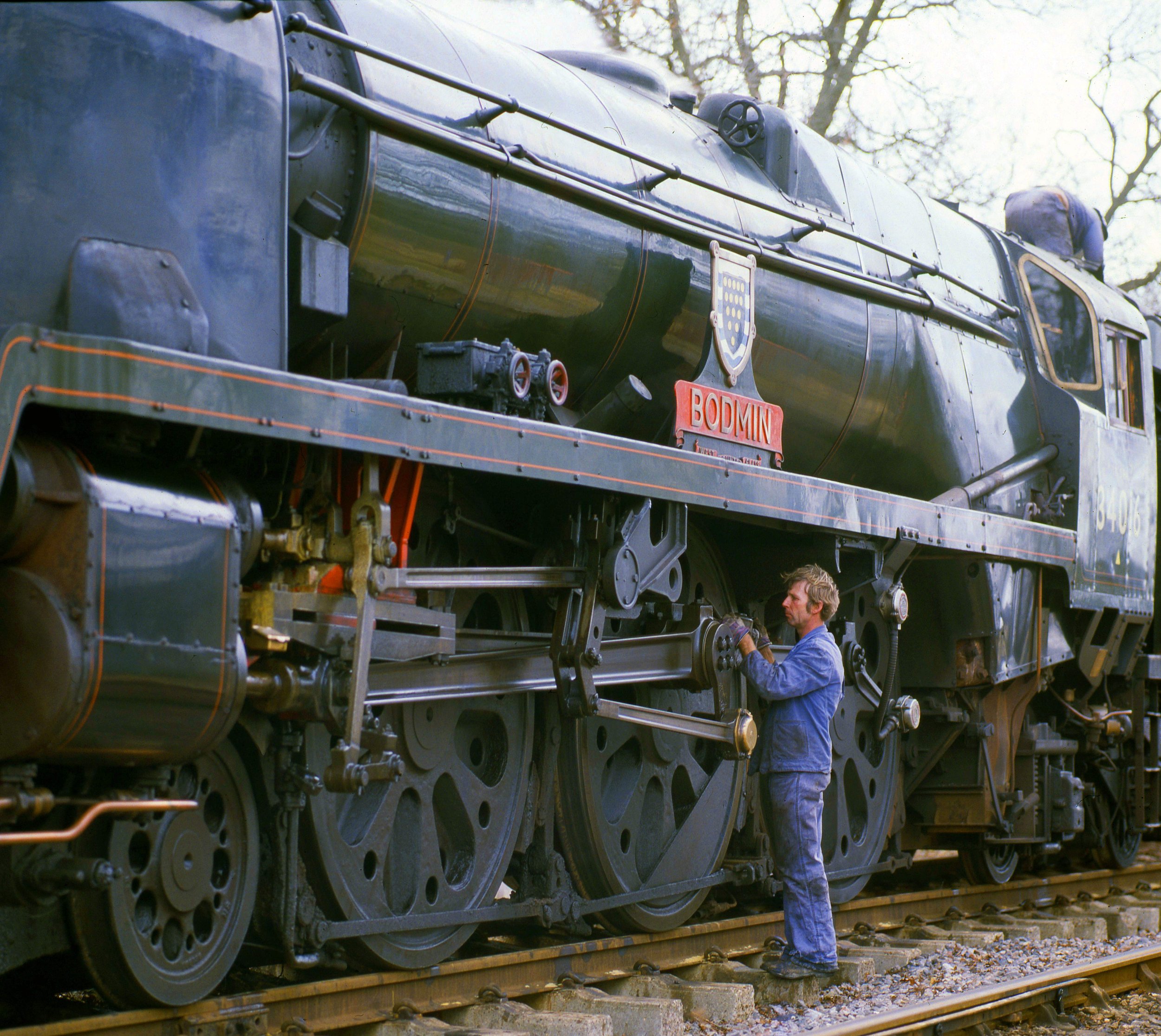 A man in a blue work jumpsuit is repairing a large black steam locomotive outdoors, with trees in the background. The train is on tracks, and the man is focused on a component near the underside of the locomotive.