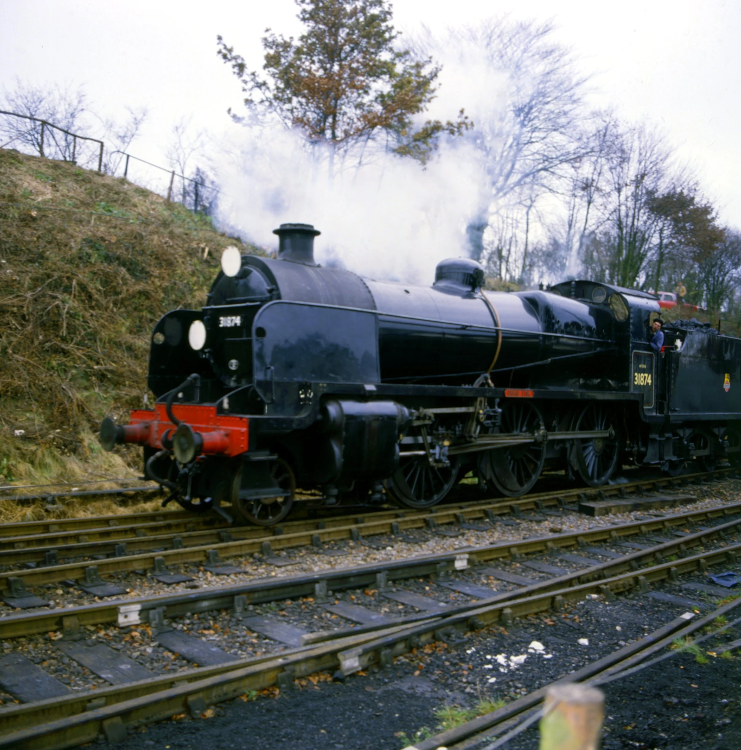 A vintage black steam locomotive on railway tracks with smoke coming from its chimney, set against a backdrop of leafless trees and cloudy sky.