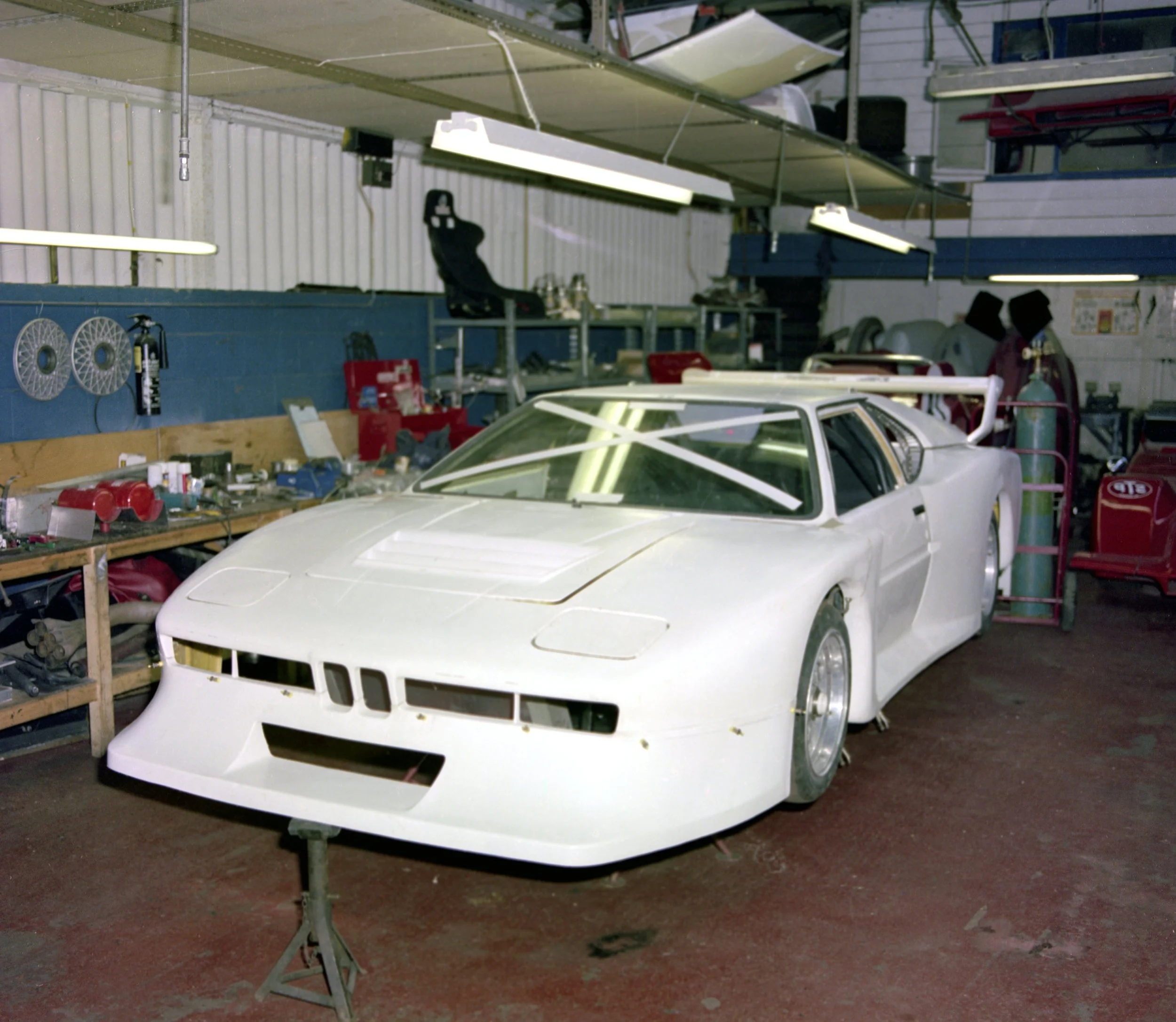 A diecast model of a white sports car with a sleek design and tinted windows, situated on a workbench in a workshop with tools and supplies around.