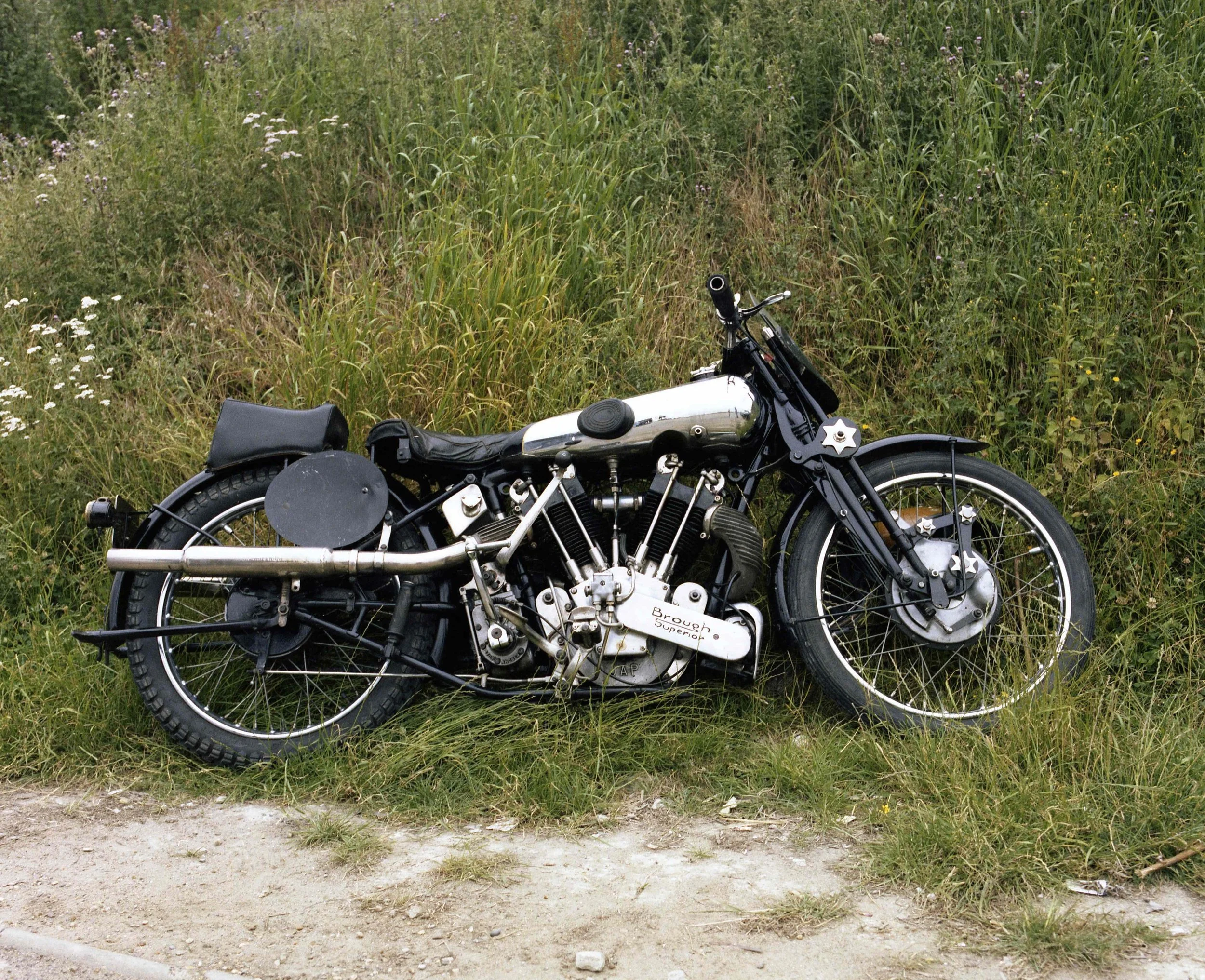 A vintage black motorcycle with a white gas tank parked on a grassy patch next to a dirt road and dense tall grass and wildflowers.