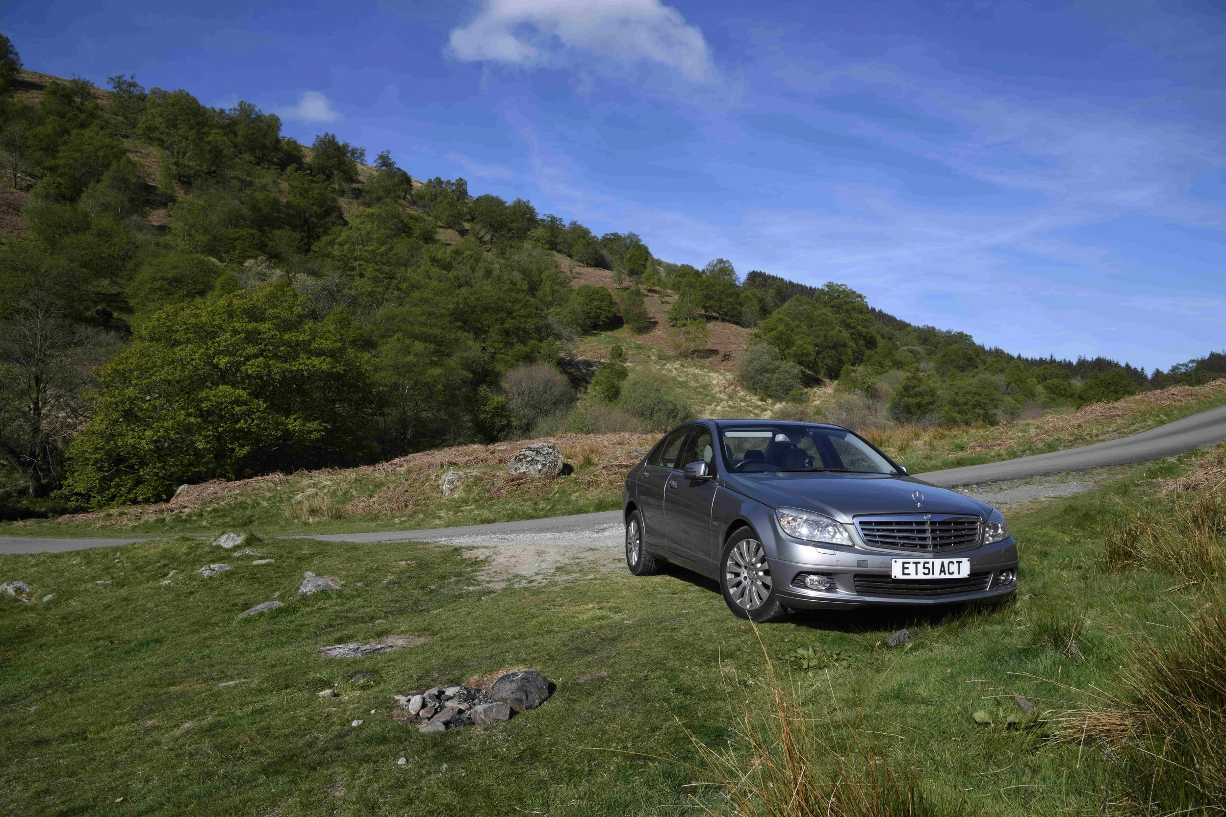 A silver Mercedes-Benz car parked on a grassy area near a winding road. Behind the car, there is a hillside covered with green trees and bushes. The sky is mostly clear with a few clouds.
