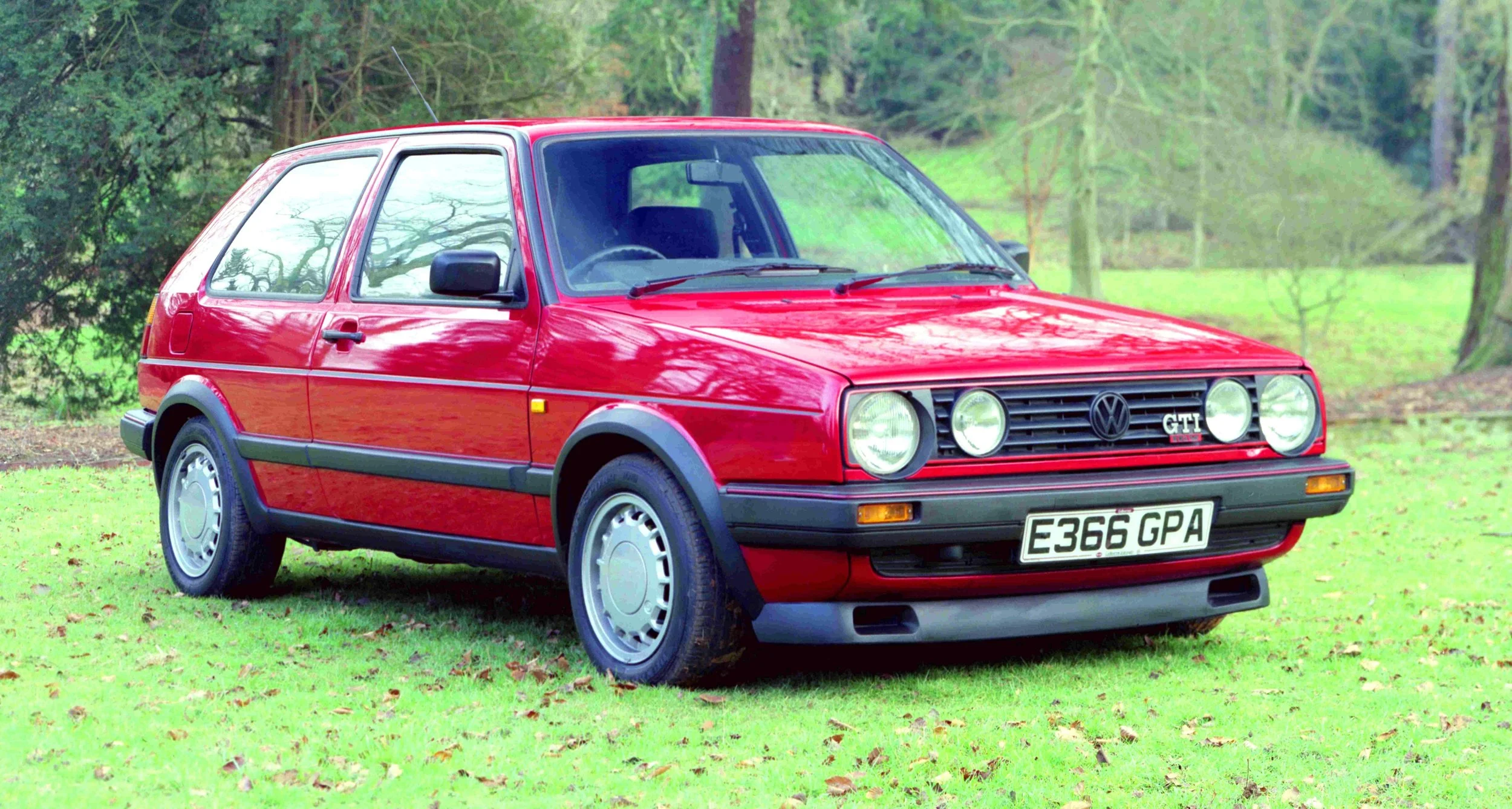 A red vintage Volkswagen GTI car parked on a grassy area with trees in the background.