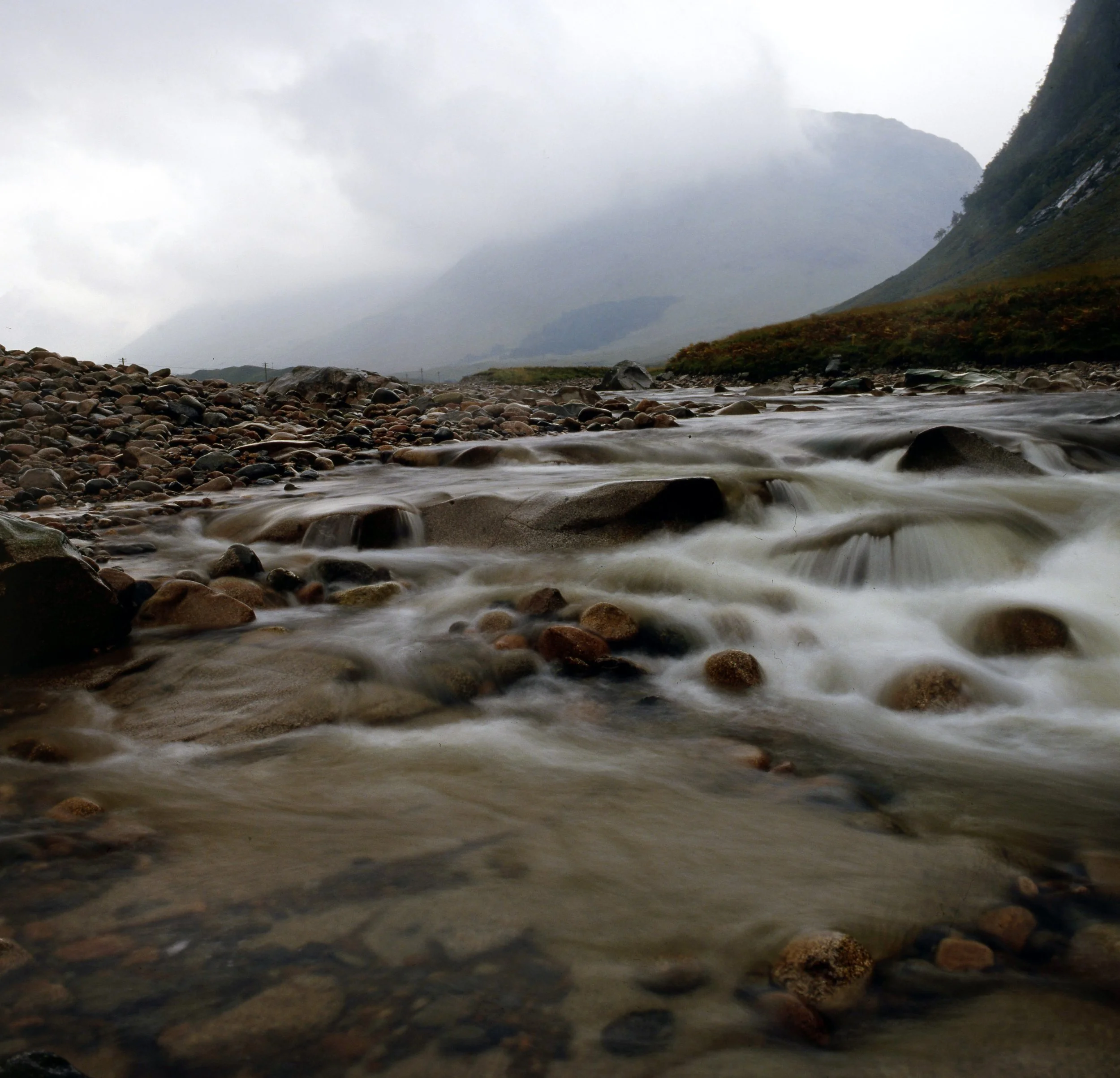 A mountain landscape with a cloudy sky, a rocky shoreline, and a flowing stream or river.