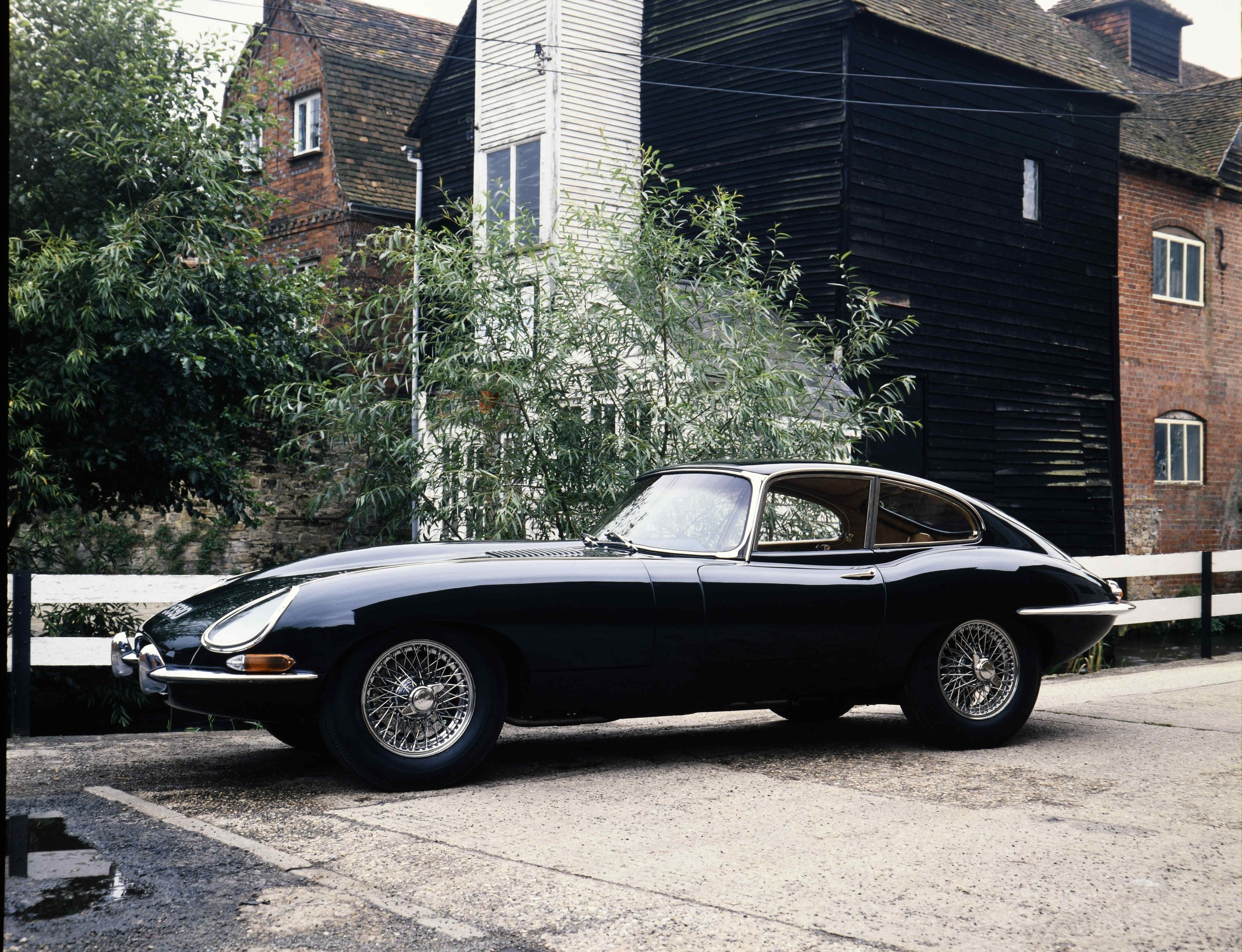 A classic black sports car parked on a street in front of old brick buildings with a wooden extension painted black and white siding, surrounded by greenery.