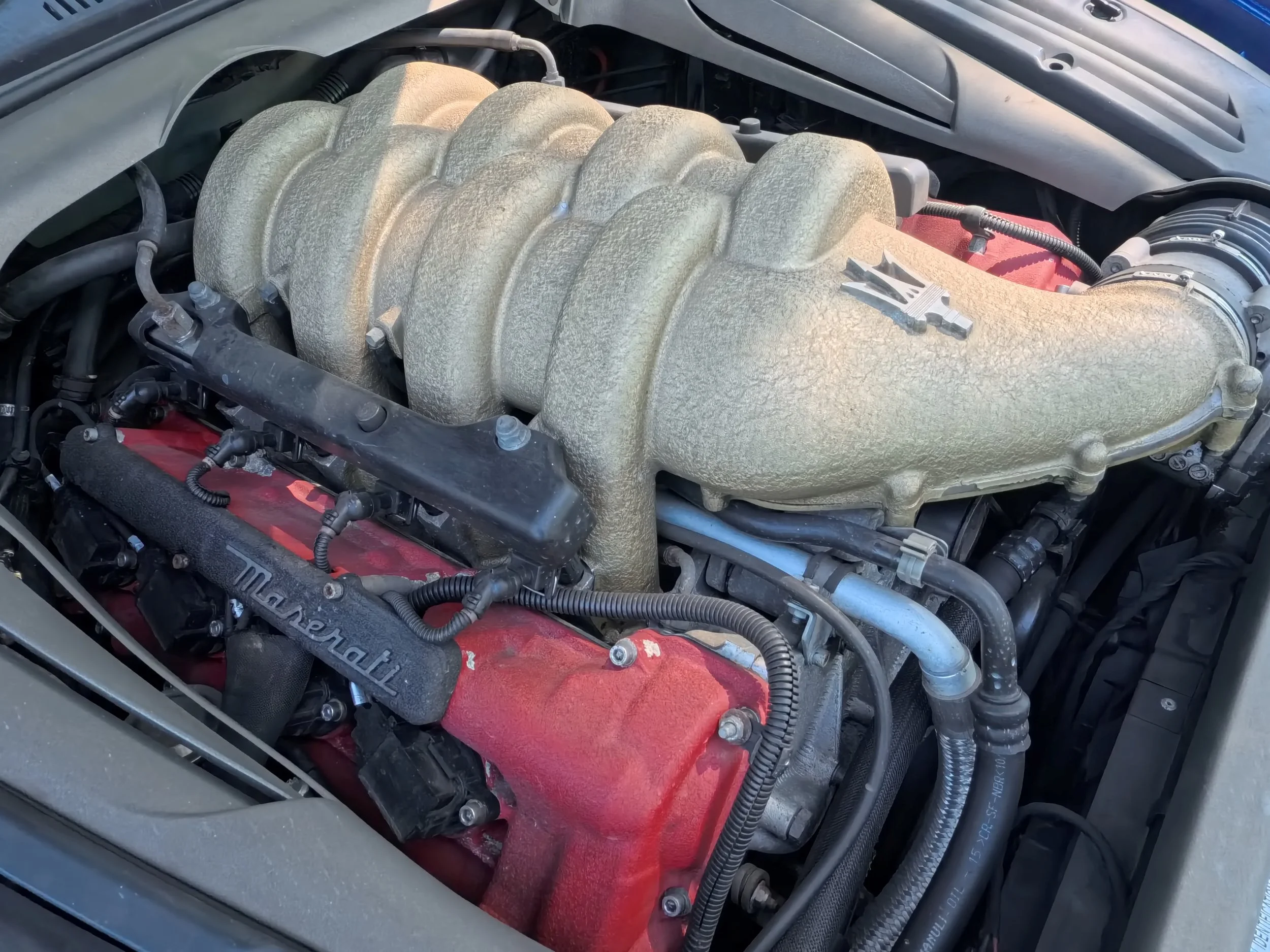 A car engine with a large, textured intake manifold in the center, various hoses, wires, and components surrounding it, including a red valve cover.