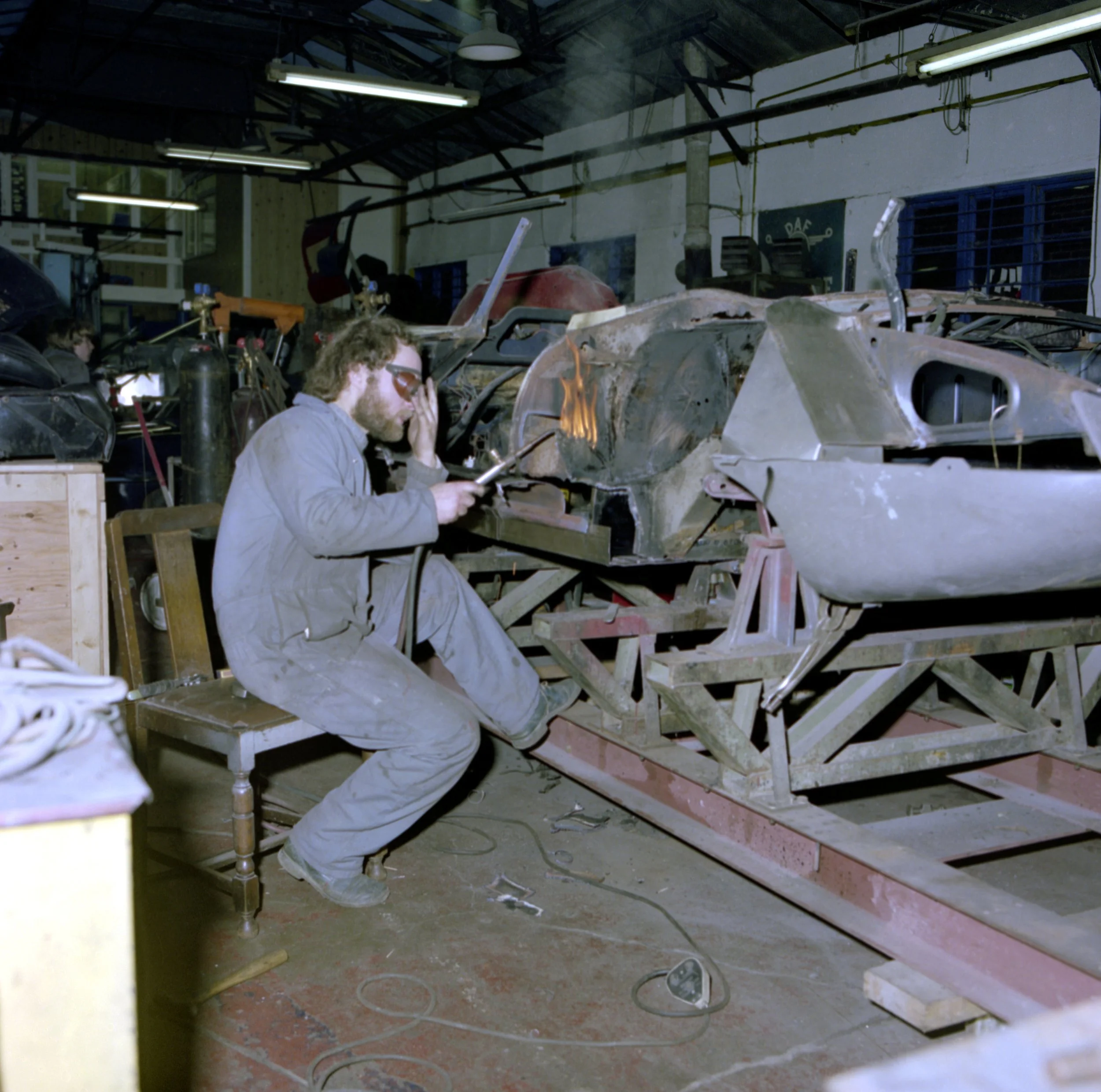 A person wearing safety goggles working on a car frame in a workshop, with various tools and equipment around, and fluorescent lights overhead.