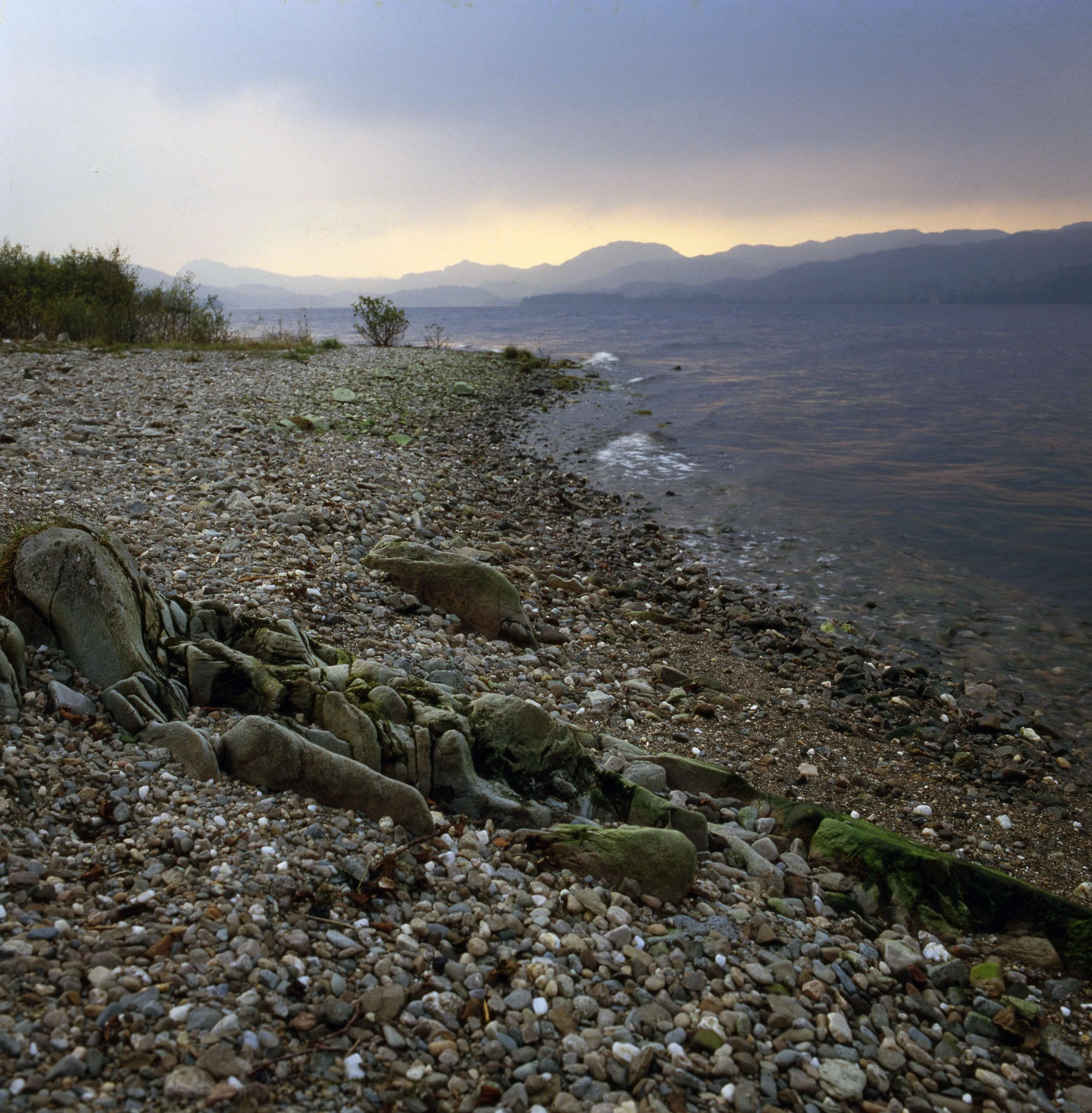 A rocky beach with small to medium-sized stones and driftwood, with a body of water and distant mountains under a cloudy sky.