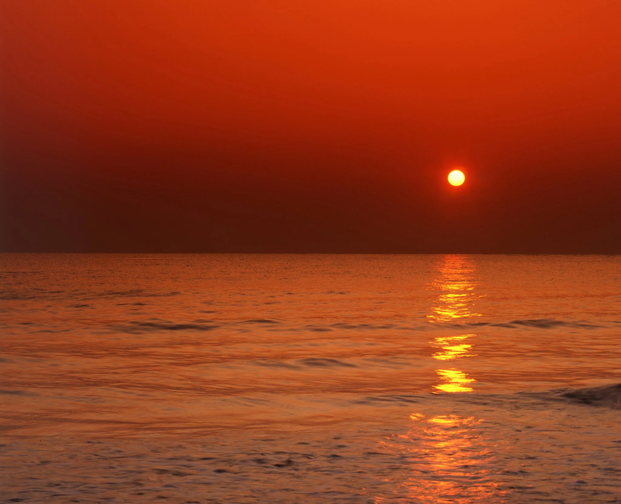 A sunset over the ocean with a small bright sun near the horizon, casting orange and red hues across the sky and reflecting on the water.