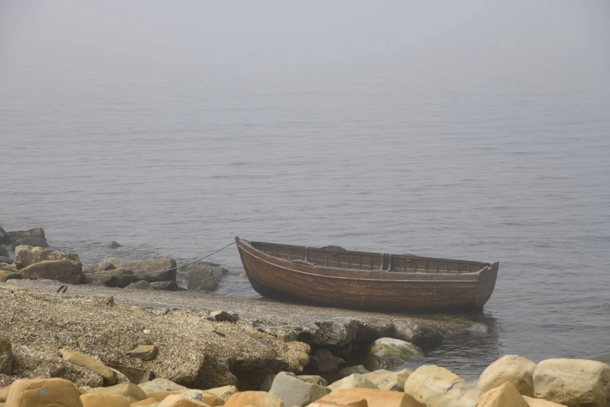 An old wooden boat resting on the rocky shore beside calm water with a misty or foggy background.