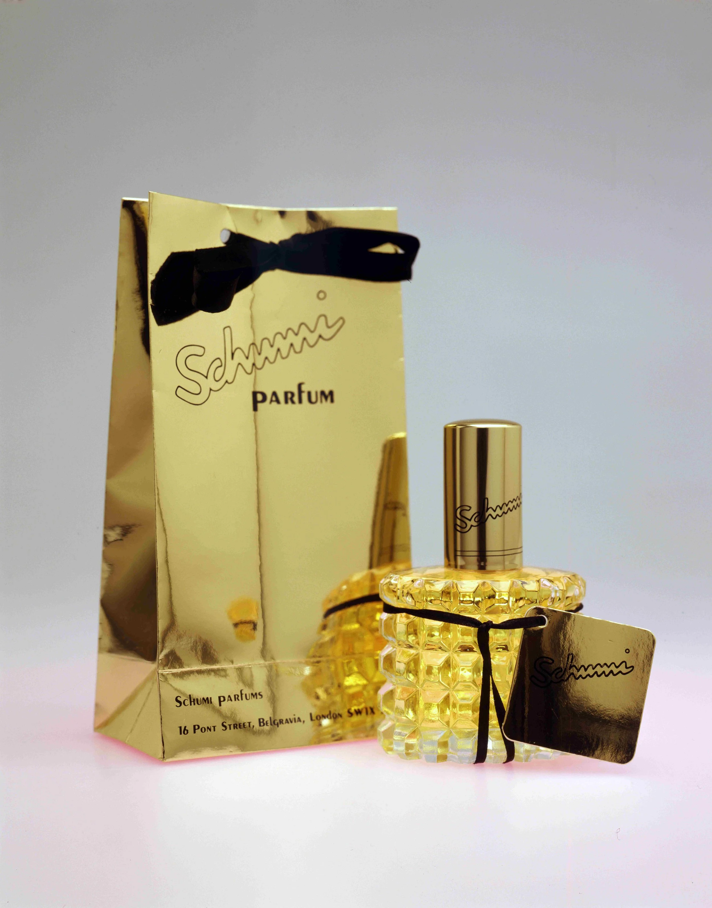 A gold-colored shopping bag with black string handles, a bottle of yellow perfume with a textured glass design, and a black tag attached with more text, all displayed against a plain white background.