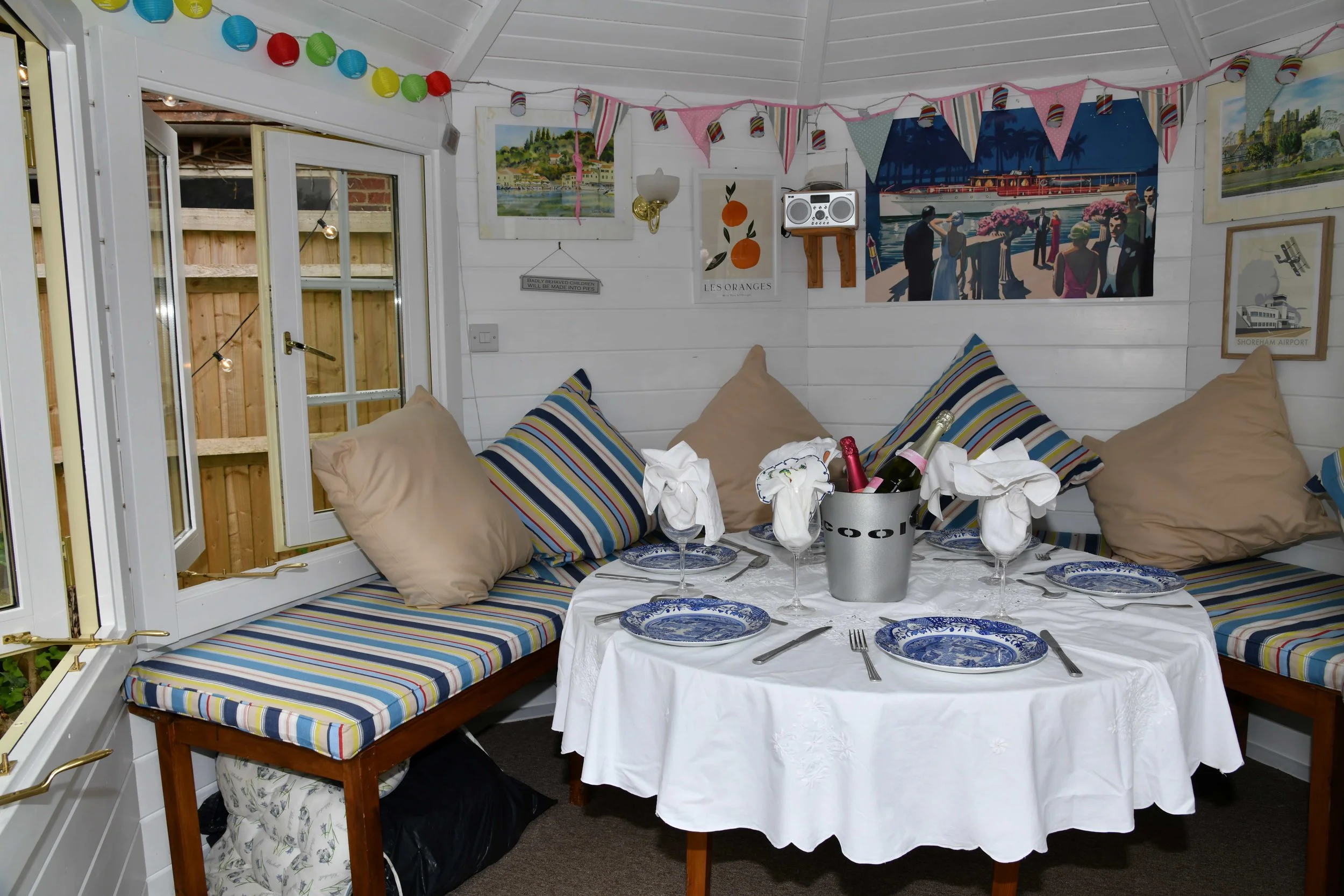 A cozy dining area decorated with colorful paper lanterns and bunting banners on the ceiling. The walls are adorned with landscape and travel-themed artwork. There is a table set with blue and white patterned plates, silverware, and wine glasses, wit