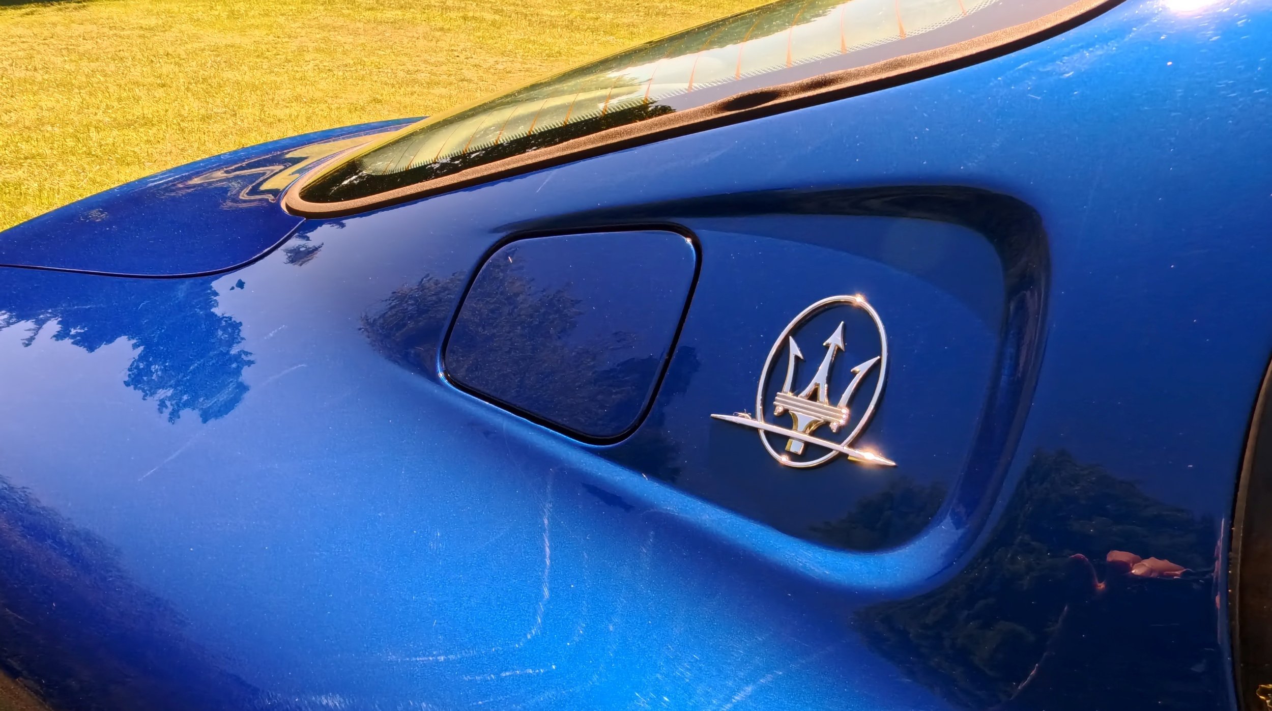 Close-up of a blue Maserati sports car with the Maserati logo on the door, parked on a grassy area.