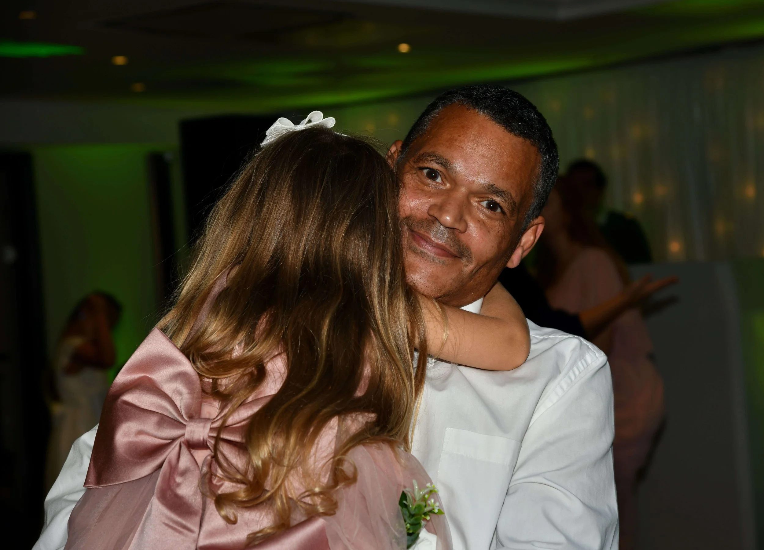 A man and a girl hugging at a celebration event. The girl has long hair with a white hair accessory and is wearing a shiny pink dress with a large bow. The man has short dark hair, is smiling, and is wearing a white shirt with a small boutonniere. Th