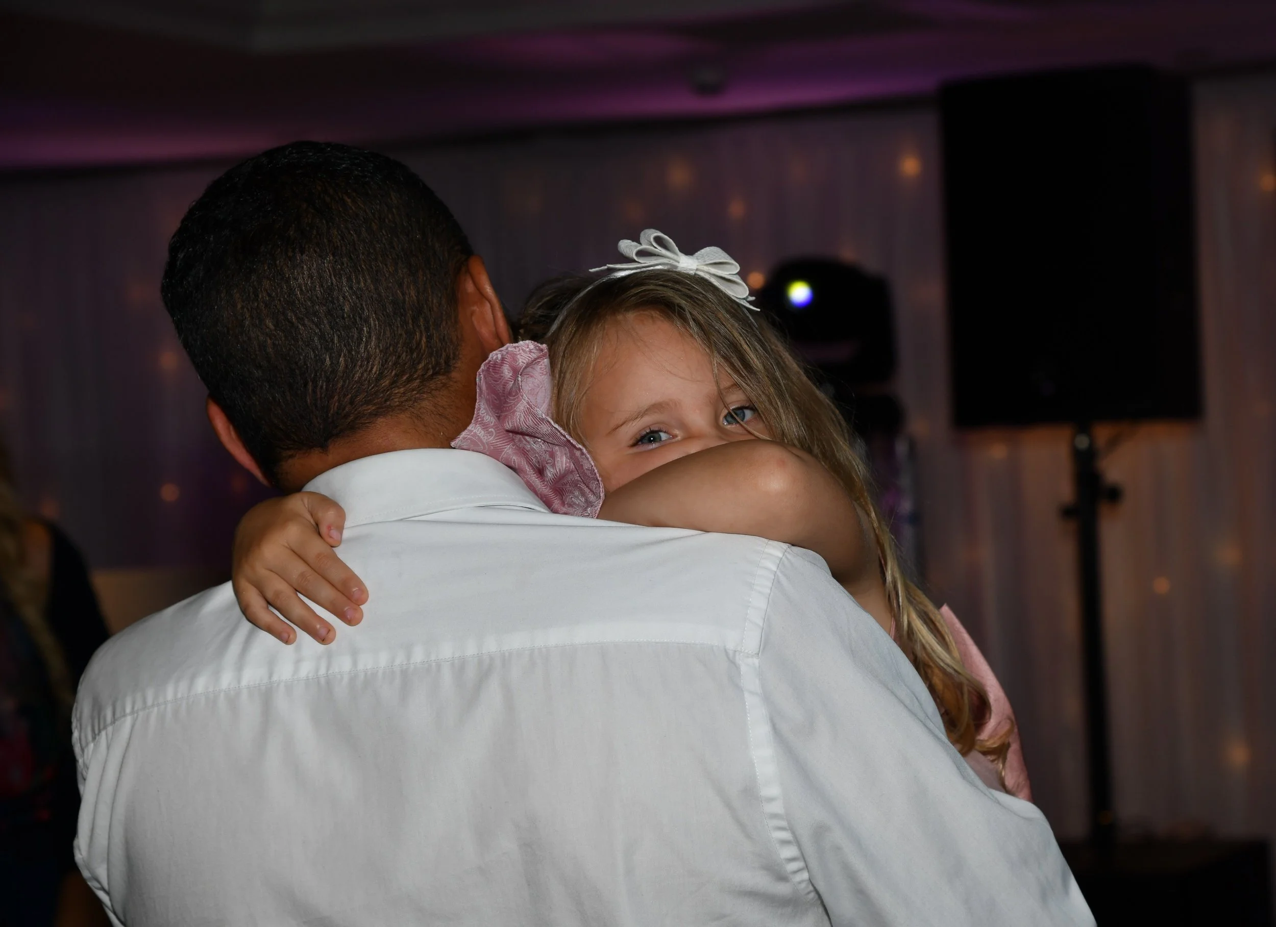 A man and a young girl are embracing. The girl is smiling and has blonde hair with a white bow, looking over the man's shoulder. The background suggests they are at a social event or party.