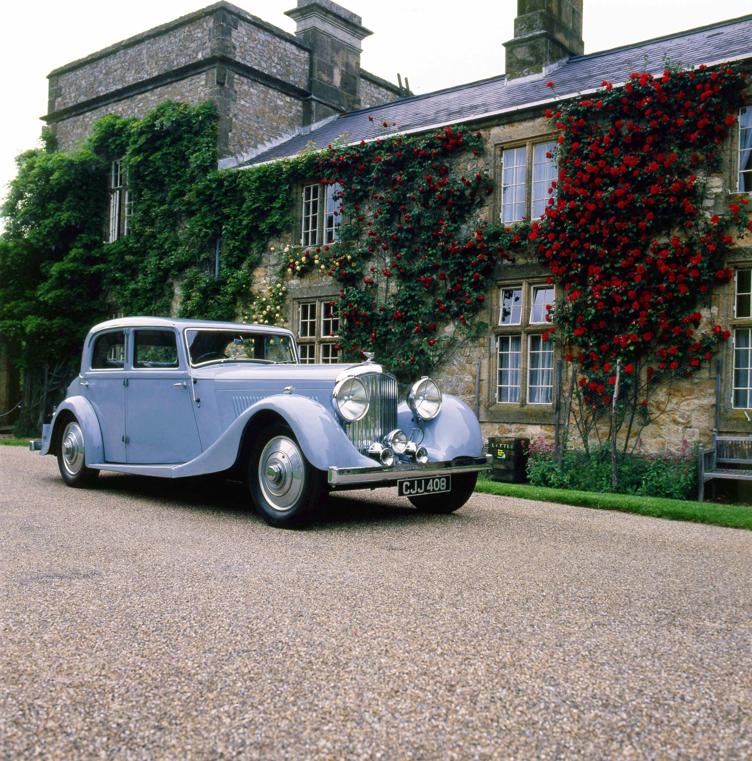 An old classic silver car parked in front of a stone house with green vines and red flowers on the facade.