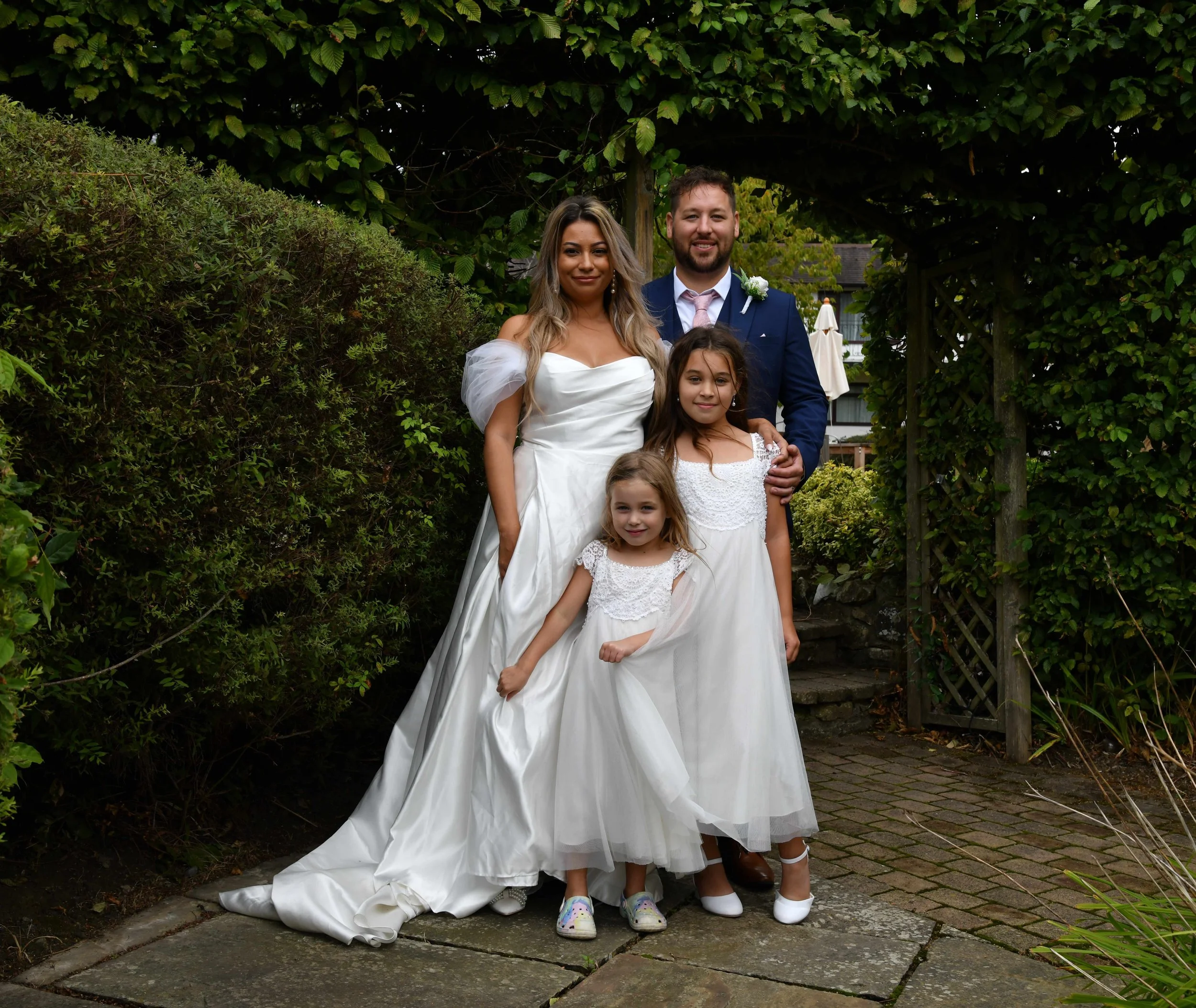 A group of five people, including a woman in a white wedding dress, two young girls in white dresses, and a man in a suit, standing outdoors on a stone patio surrounded by greenery, possibly at a wedding or special event.