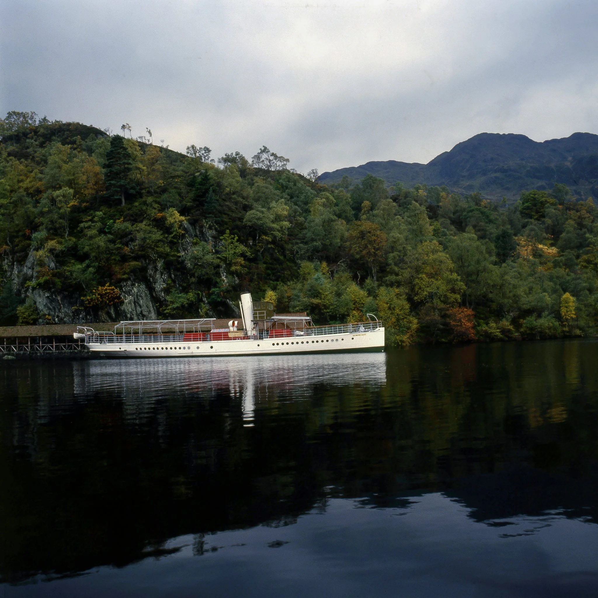 A scenic view of a boat on a calm river, with lush green trees and mountains in the background, under a cloudy sky.
