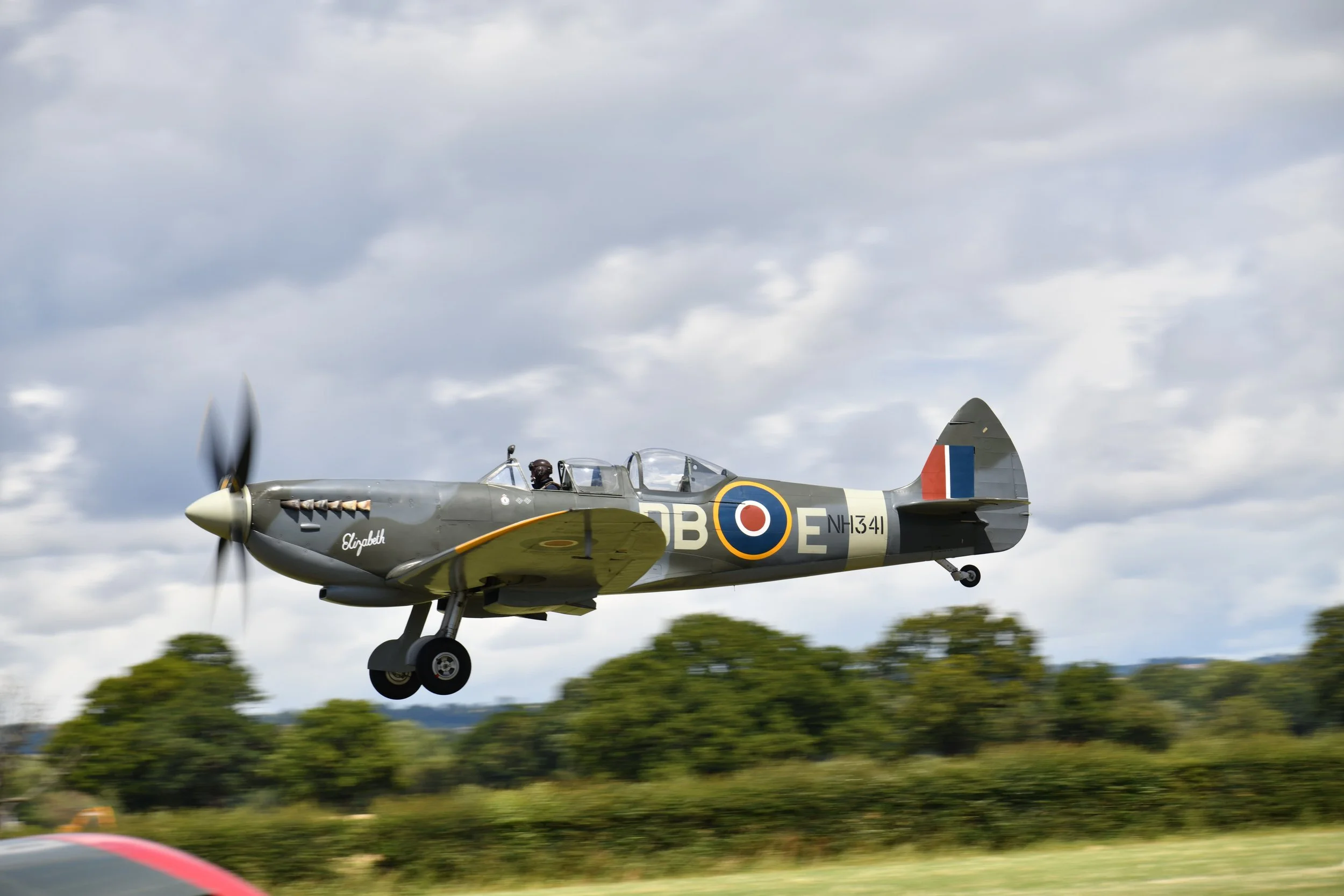 A vintage military aircraft, labeled 'Elizabeth' on the nose, flying with landing gear extended against a cloudy sky.