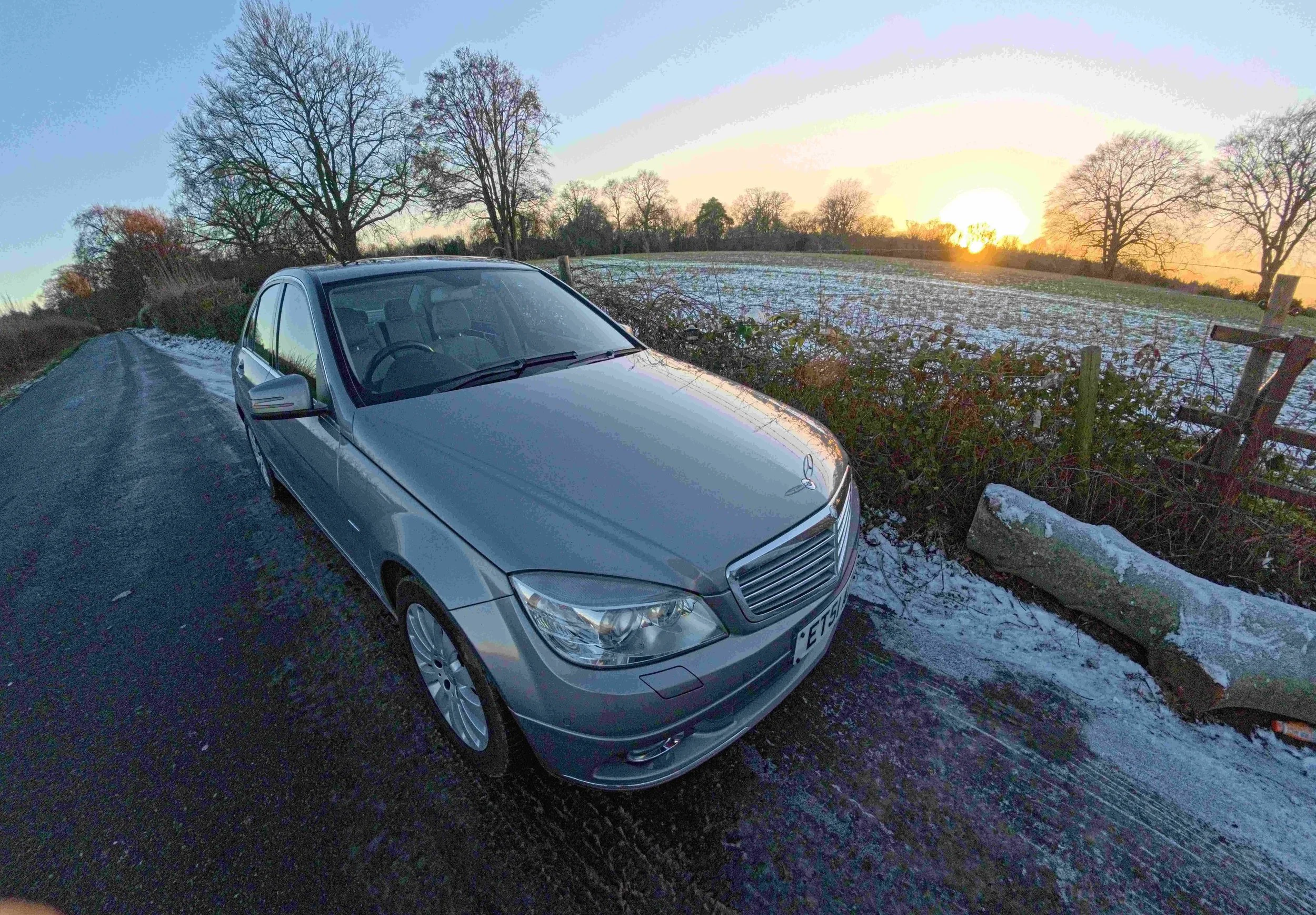 A silver Mercedes-Benz car parked on a narrow, icy country road during sunset, with leafless trees and a snow-covered field