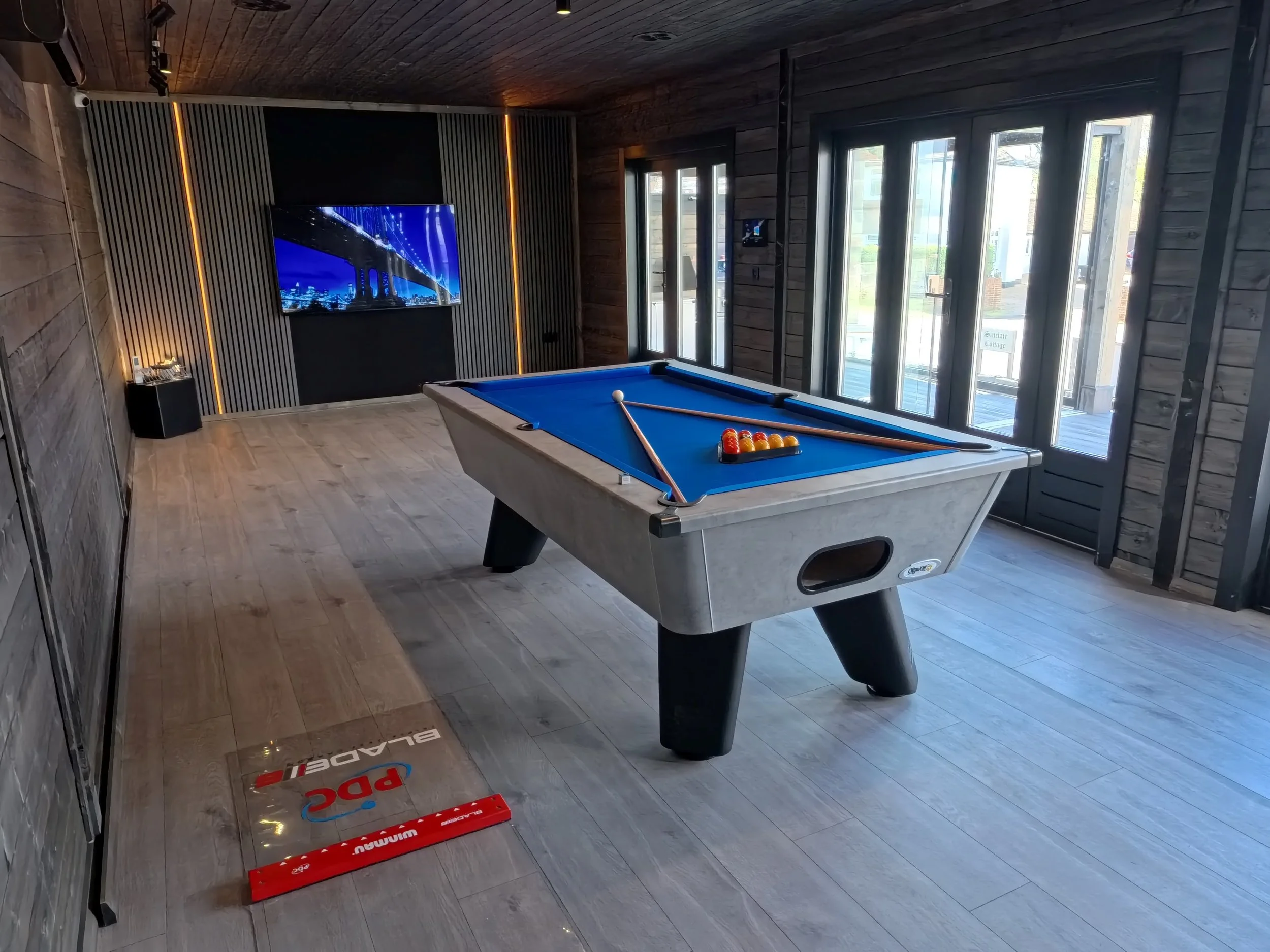 A modern living room with wood-paneled walls, a large flat-screen TV mounted on the wall, a pool table with a cue stick and balls, large floor-to-ceiling windows, and a hardwood floor.