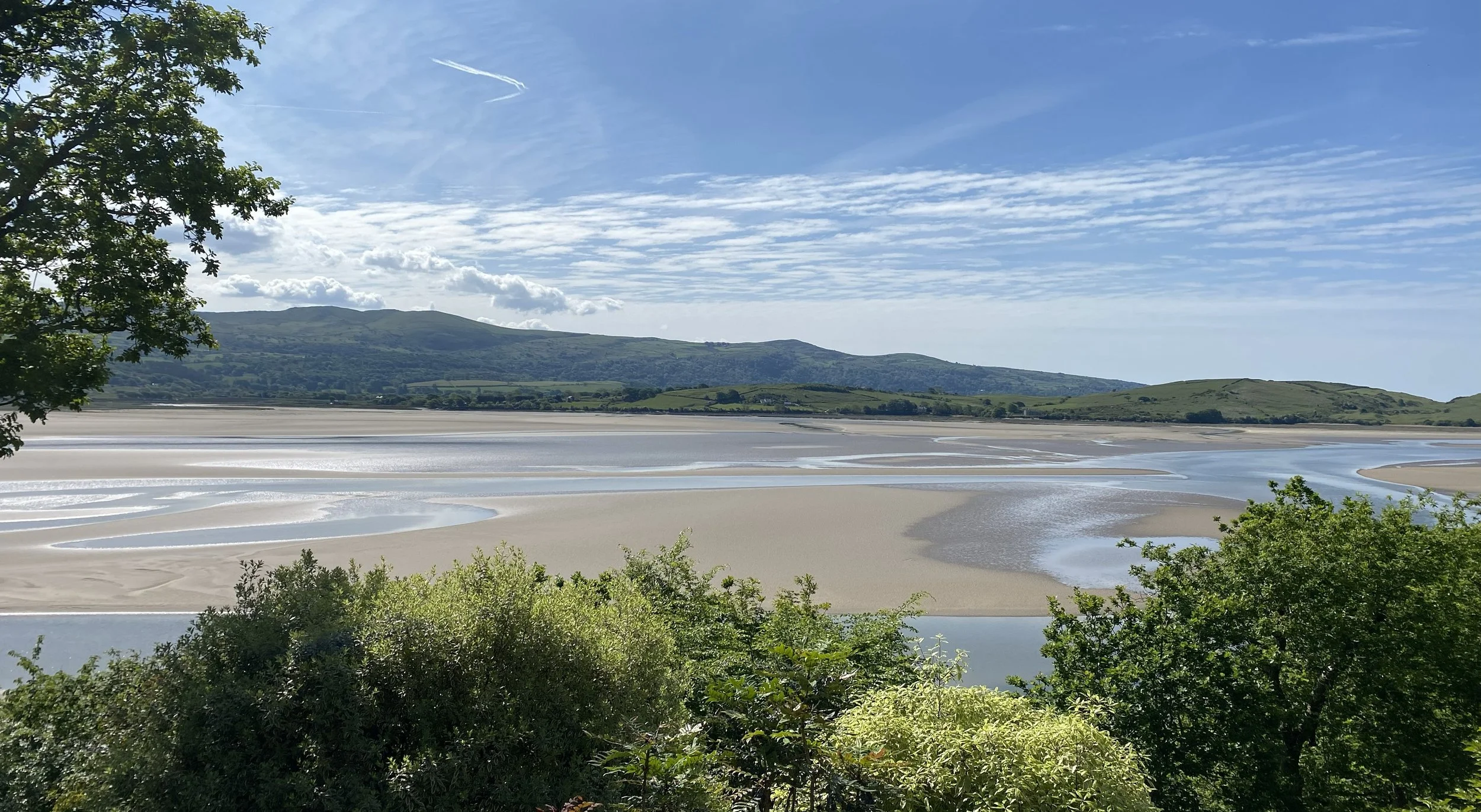 Scenic landscape of a sandy beach with winding water channels, green bushes in the foreground, and hills with a partly cloudy sky in the background.