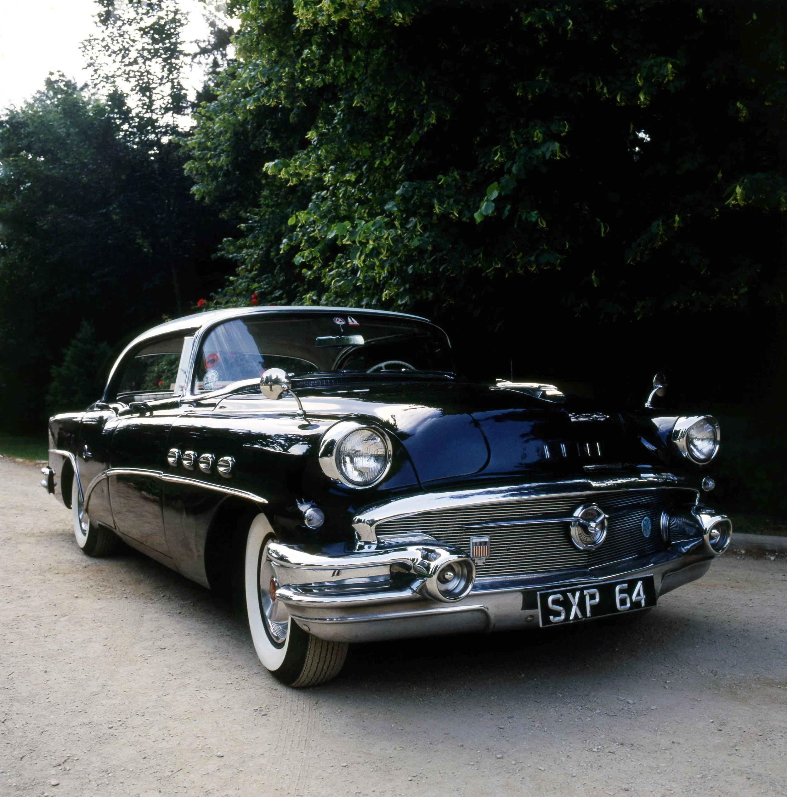 A classic black vintage car with white tires parked on a dirt surface, with dense green trees in the background.