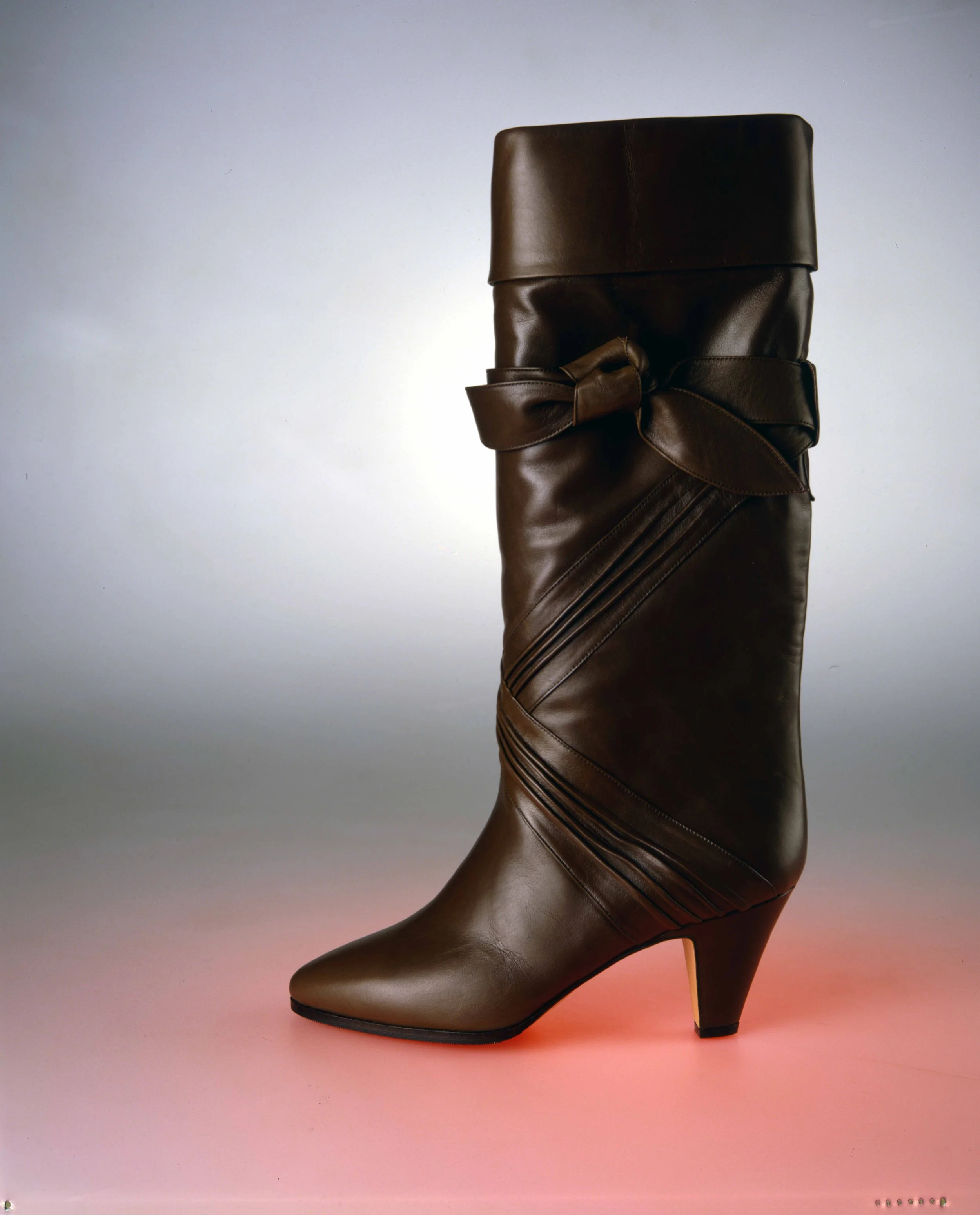 A tall, brown leather high-heeled boot with a bow on the side, set against a gradient background with warm pink and cool gray tones.
