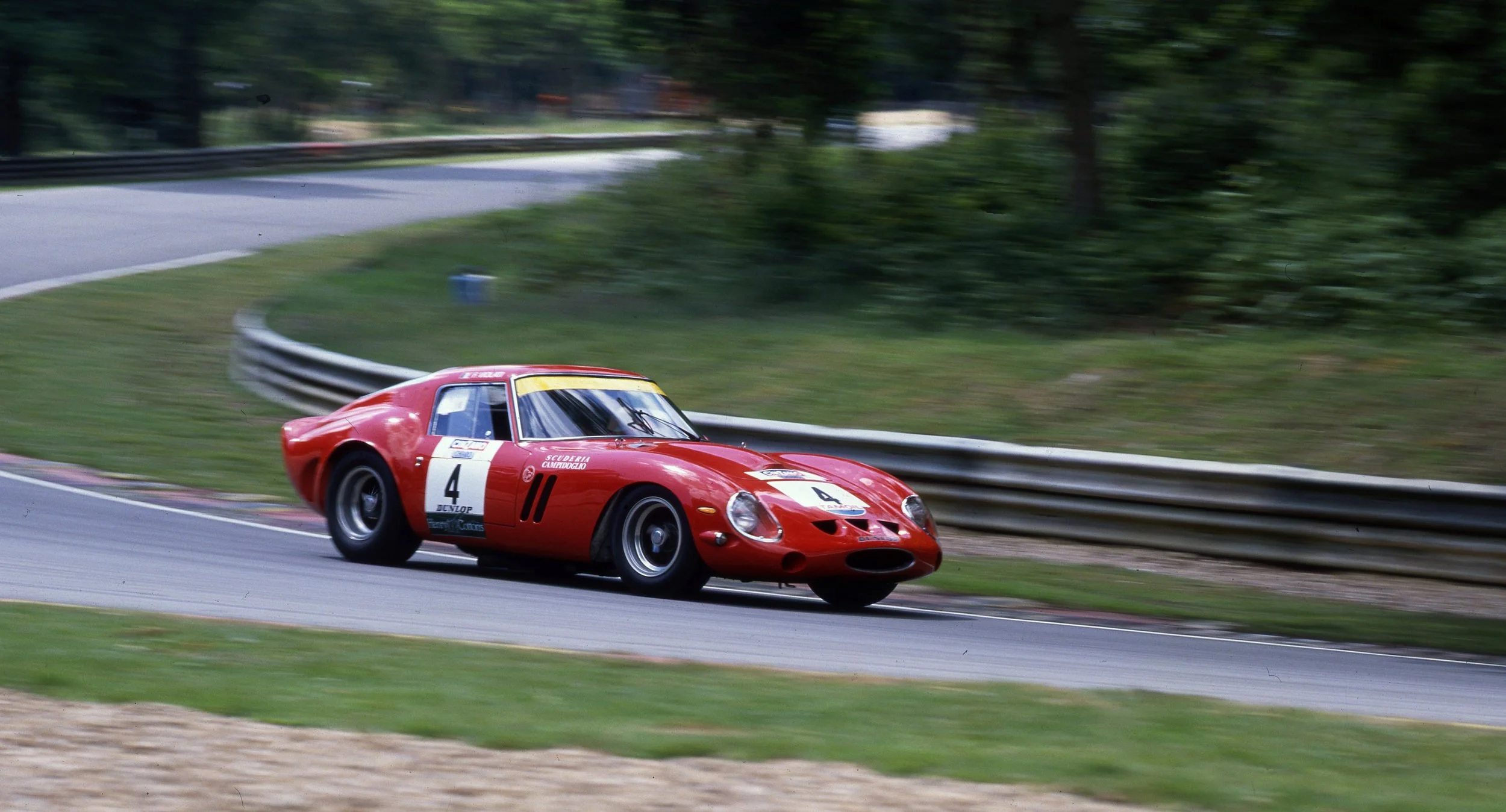 A red vintage race car with the number 4 on its side speeding around a curve on a race track, with blurred trees and guardrails in the background.