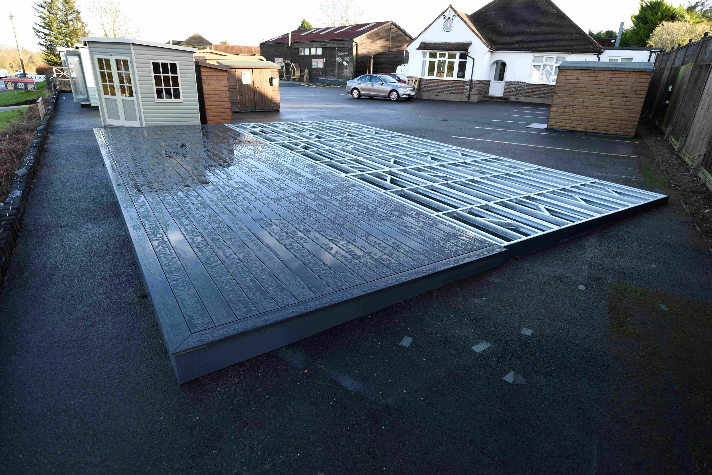 A parking lot with a partially installed metal modular wheelchair lift platform with a wood-patterned surface, surrounded by small sheds and adjacent to a building with white walls and large windows.