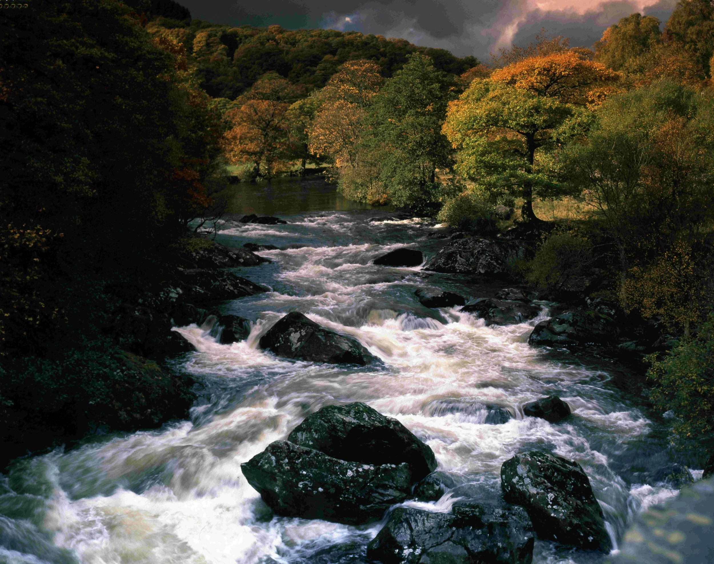 A flowing river surrounded by dense forest with autumn-colored foliage, dark clouds overhead.
