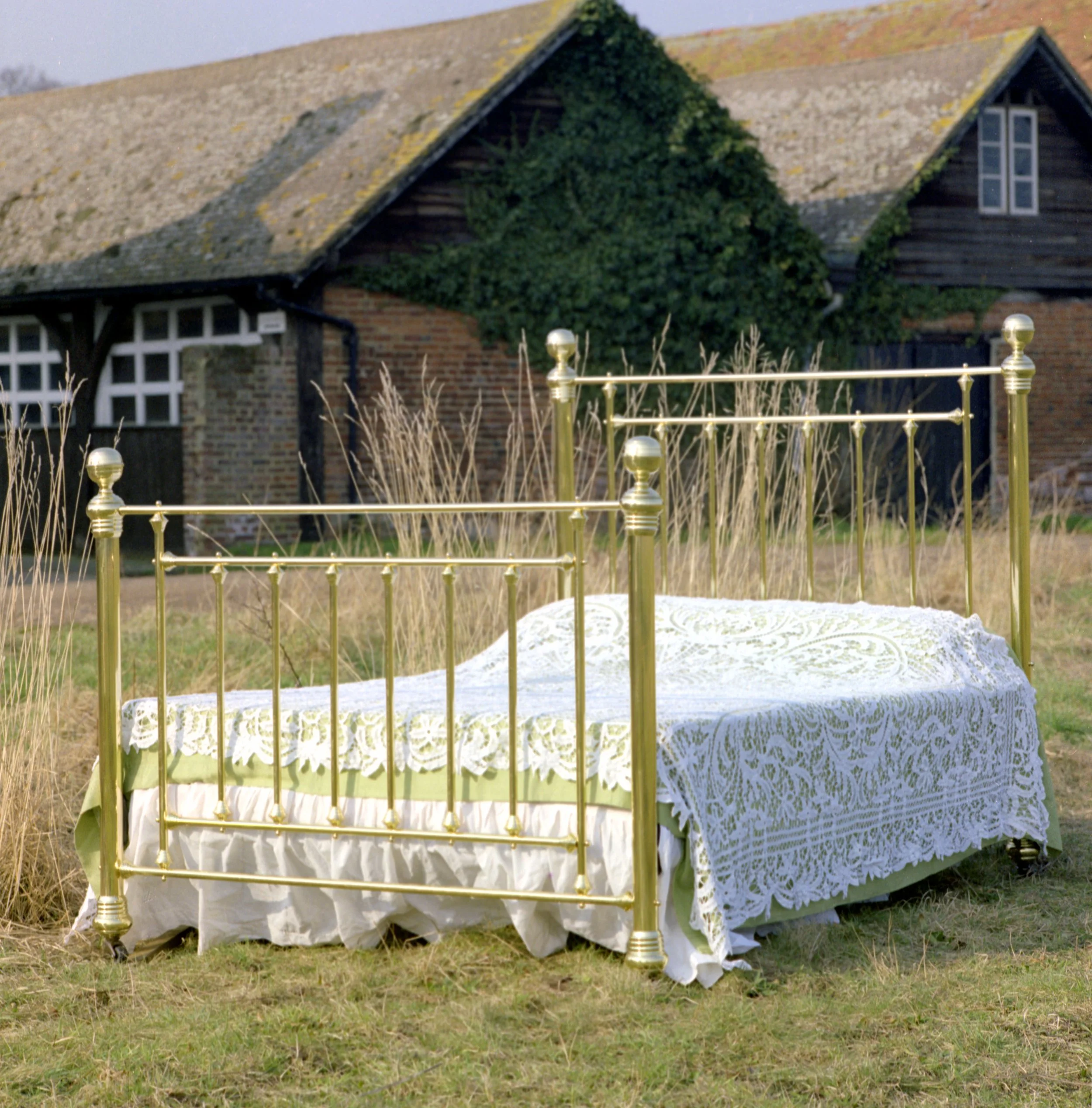 A decorative outdoor table with a gold-colored metal frame and a white lace tablecloth, positioned in front of a house with ivy-covered walls and a brick chimney, on a grassy yard.
