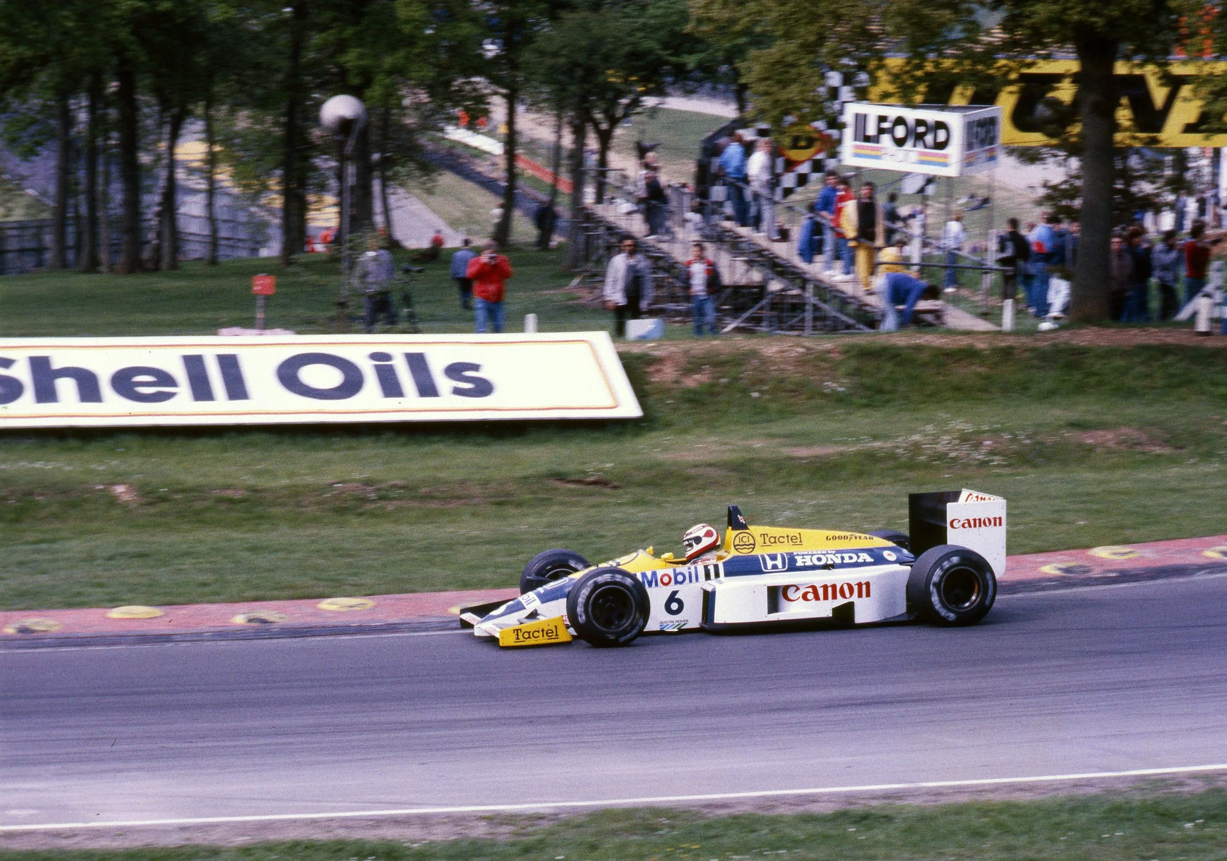 A yellow race car speeding on a track with company logos, with spectators and a scoreboard in the background.