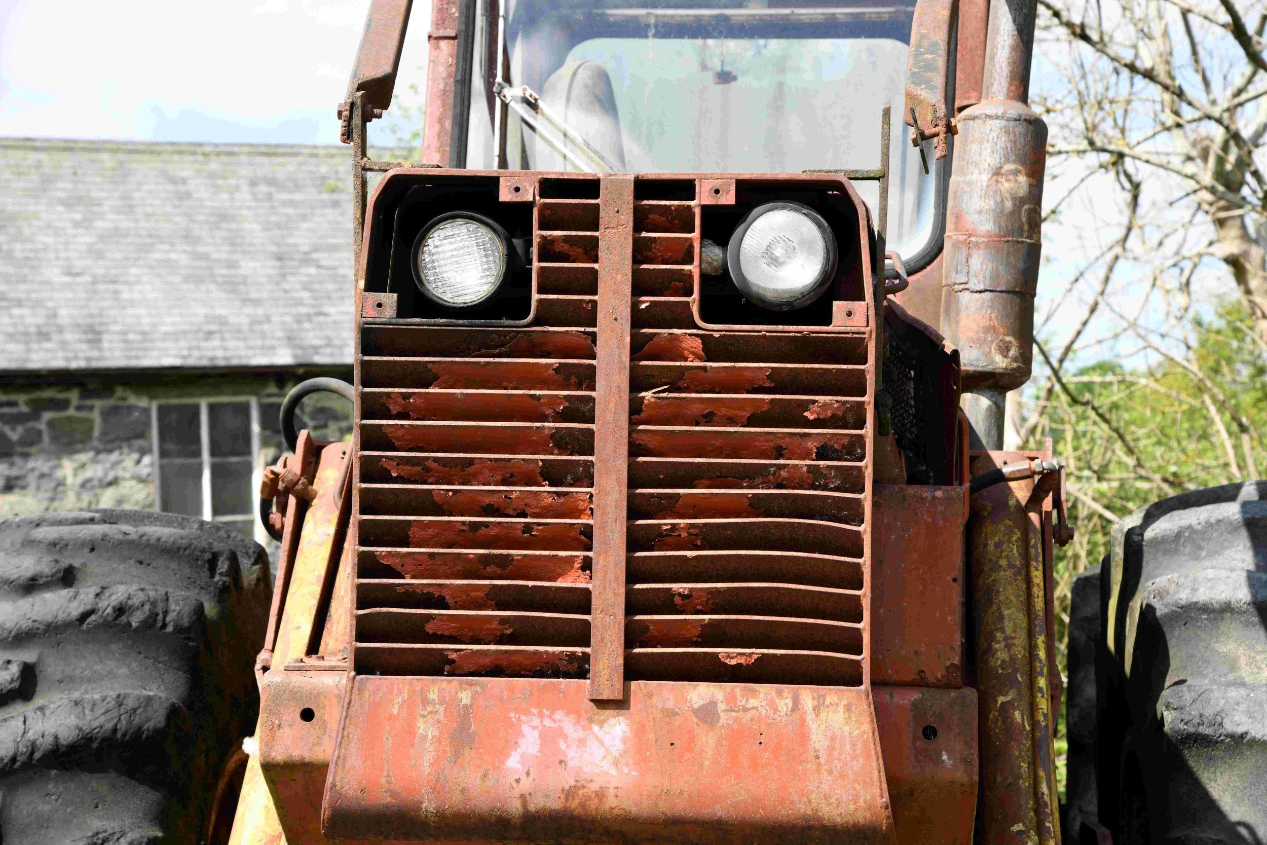 Front view of old, rusty construction vehicle with headlights and grille, outdoors with trees and a building in background.