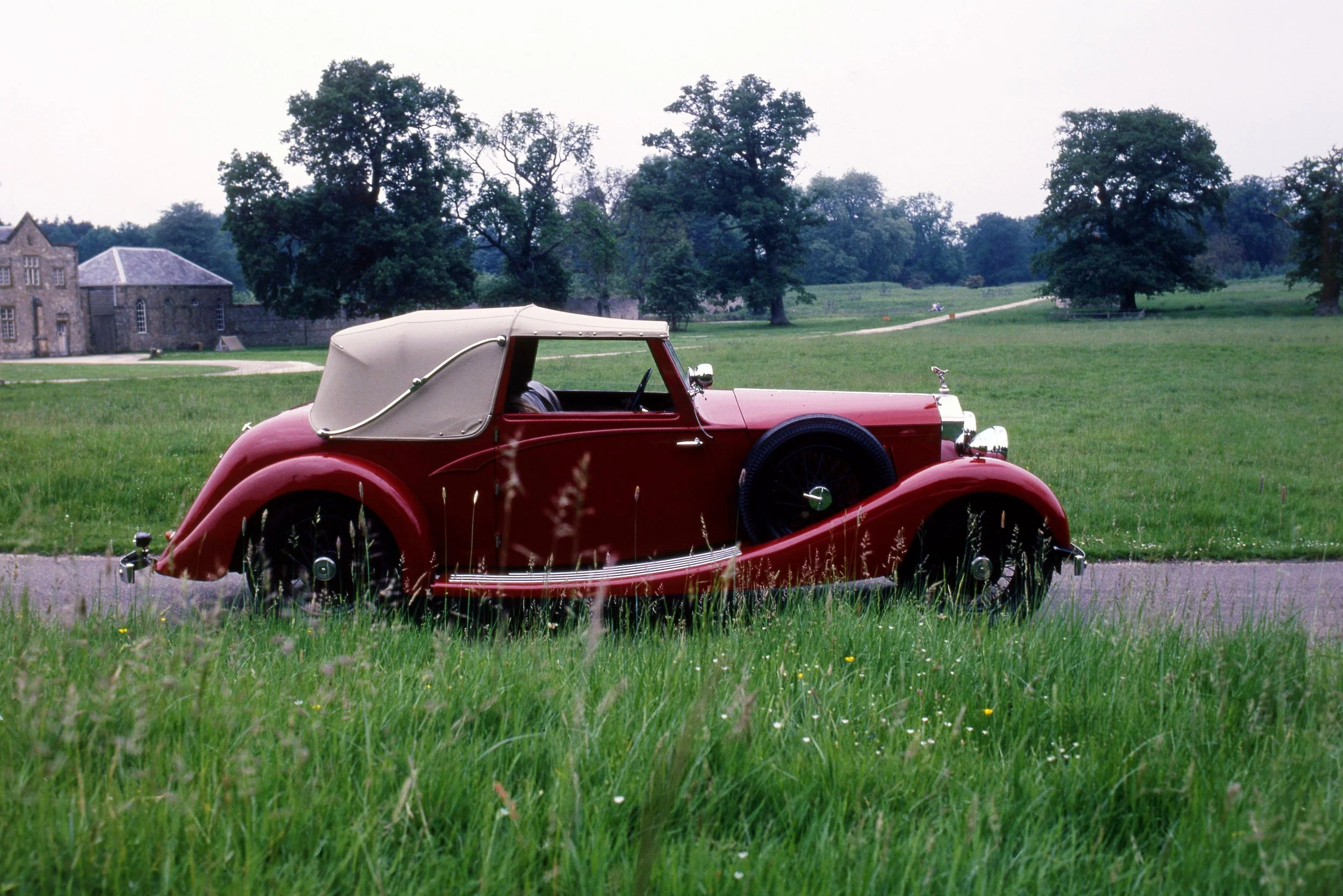 A vintage red convertible car with a beige soft top parked on a grassy field with trees and old stone buildings in the background.