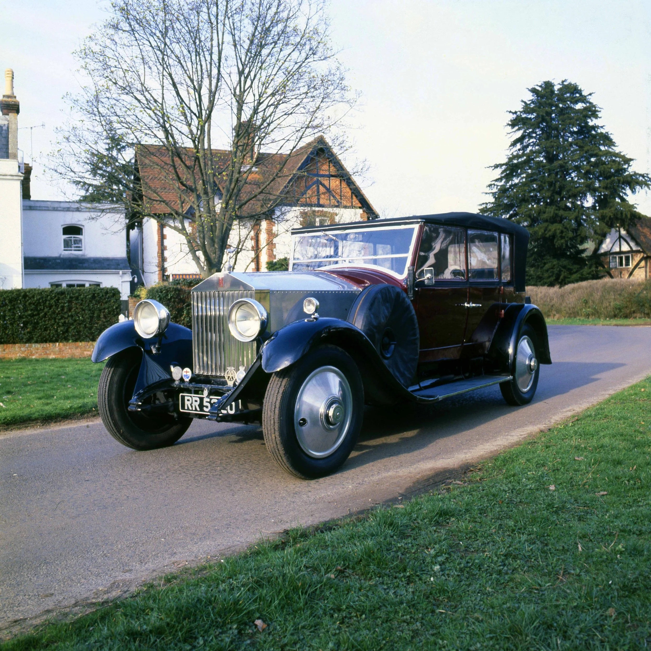 A vintage black car parked on a grassy area in front of houses with trees and a sidewalk, under a light blue sky.