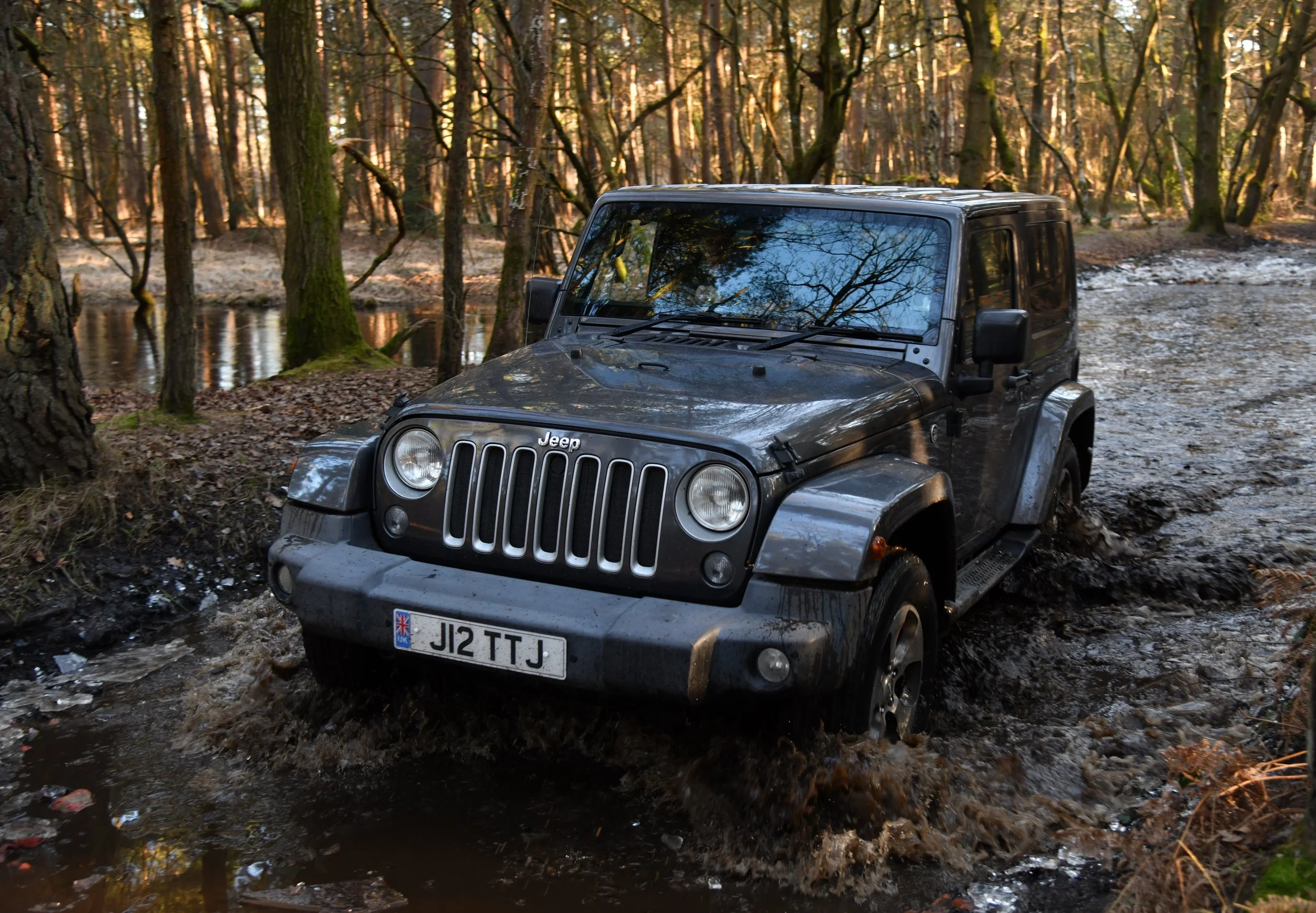 A black Jeep off-road vehicle stuck in muddy water in a forest with trees and fallen leaves.