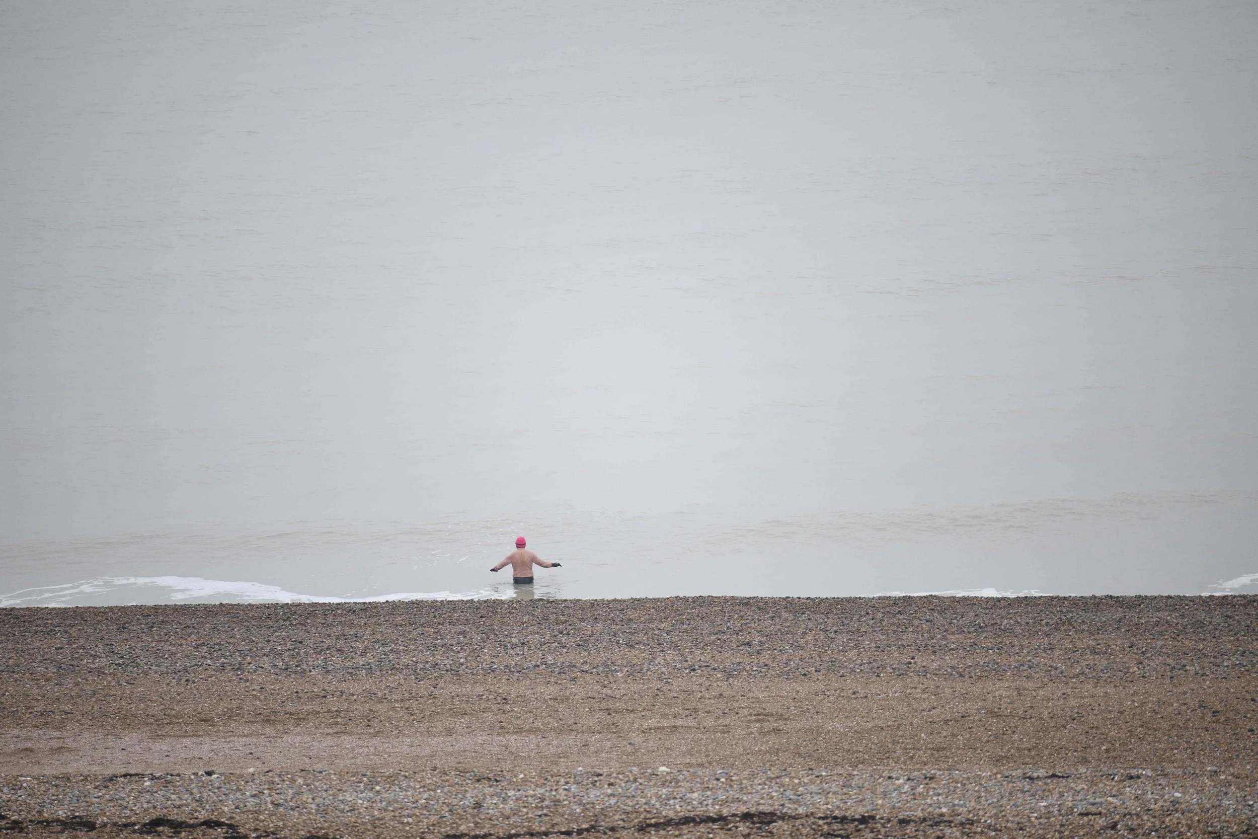 A person swimming in the ocean near the shore, wearing a pink swim cap and black swim goggles, with a cloudy sky and quiet water in the background.