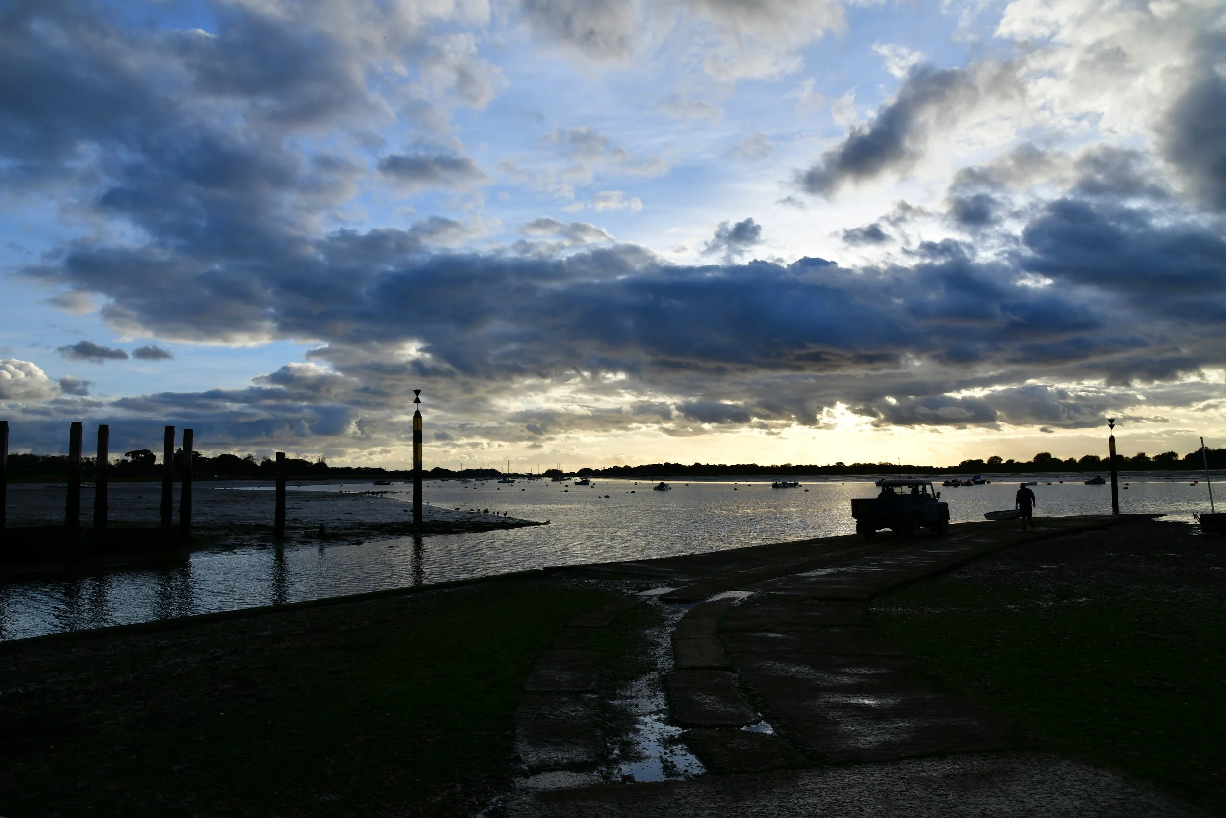 A waterfront scene during dusk or dawn with dark clouds in the sky, some boats on the water, and a person standing near a small boat on the shore, with several posts along the water's edge.