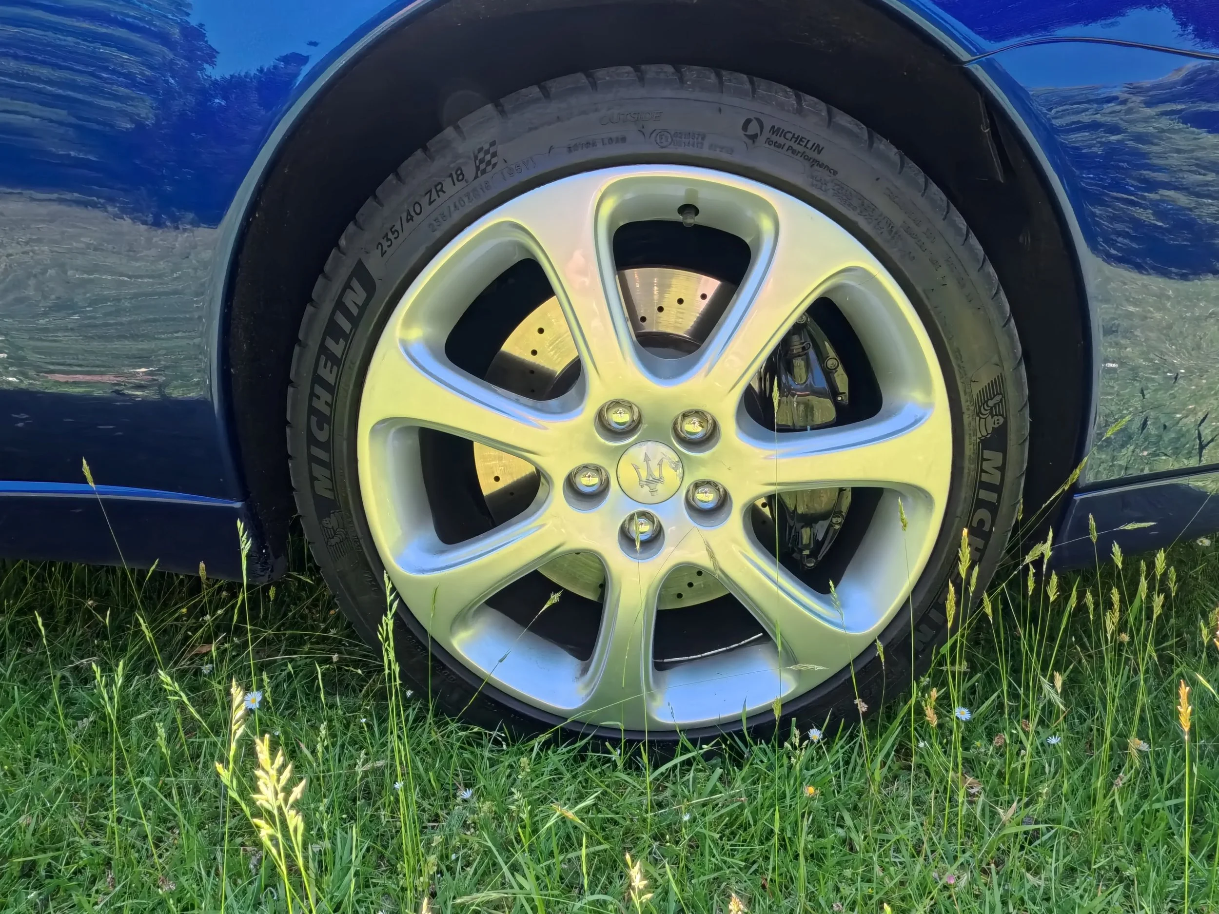 Close-up of a car wheel with a Maserati logo on the hubcap, grass and small flowers in the foreground, and a grassy area in the background.