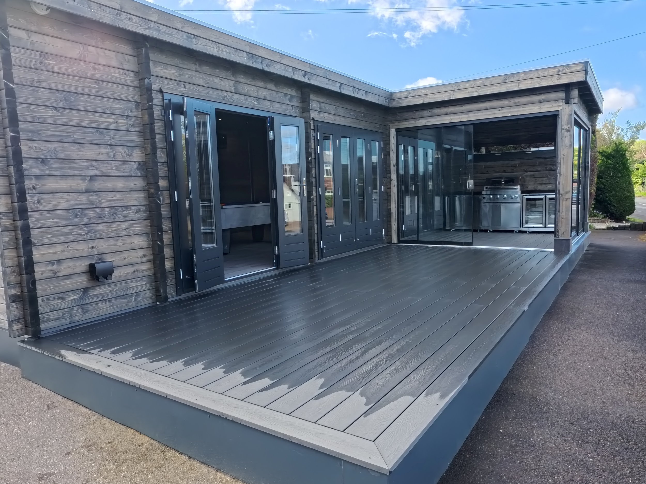 A modern outdoor kitchen and patio area with wooden walls, black framed glass doors, and stainless steel appliances, including a grill and small fridge, on a dark wooden deck under a blue sky with some clouds.