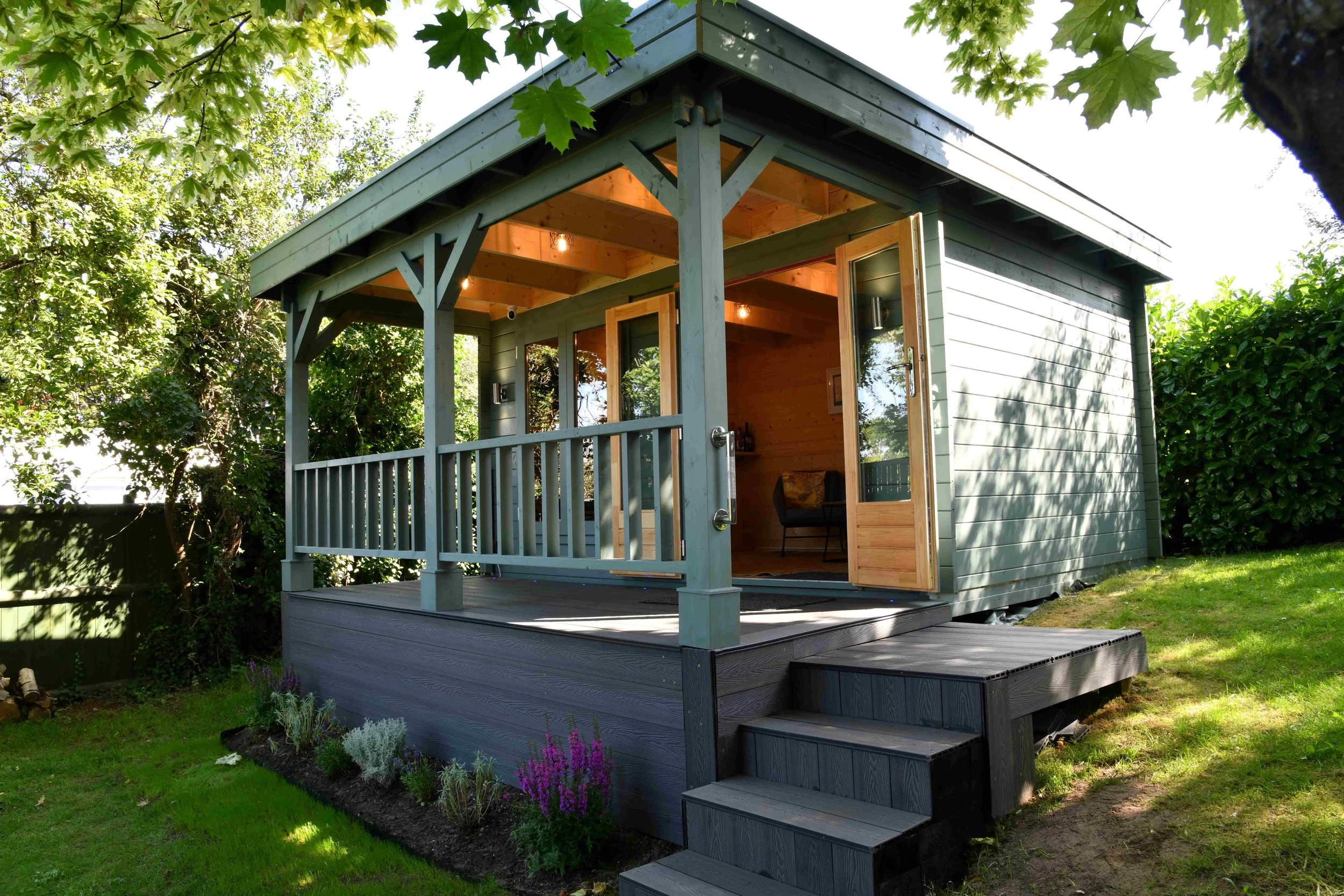 A small, modern blue house with an open door, surrounded by green trees and plants, and a small porch with white railings.