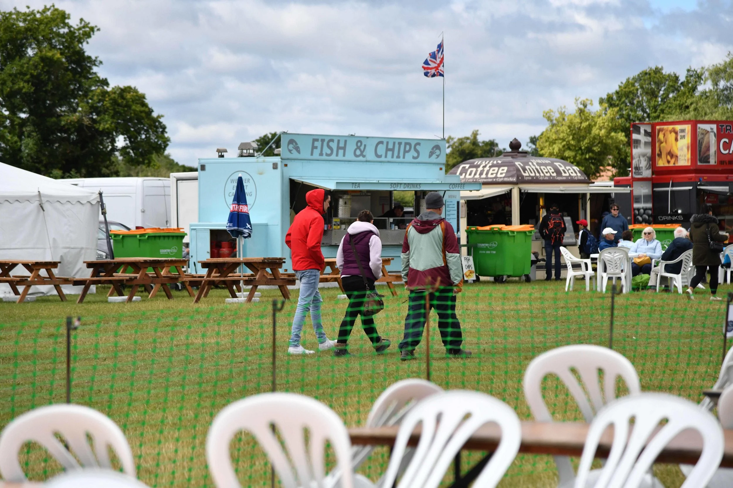 People walking near food trucks and seating area at an outdoor event, with trees, a cloudy sky, and a fenced area in the foreground.