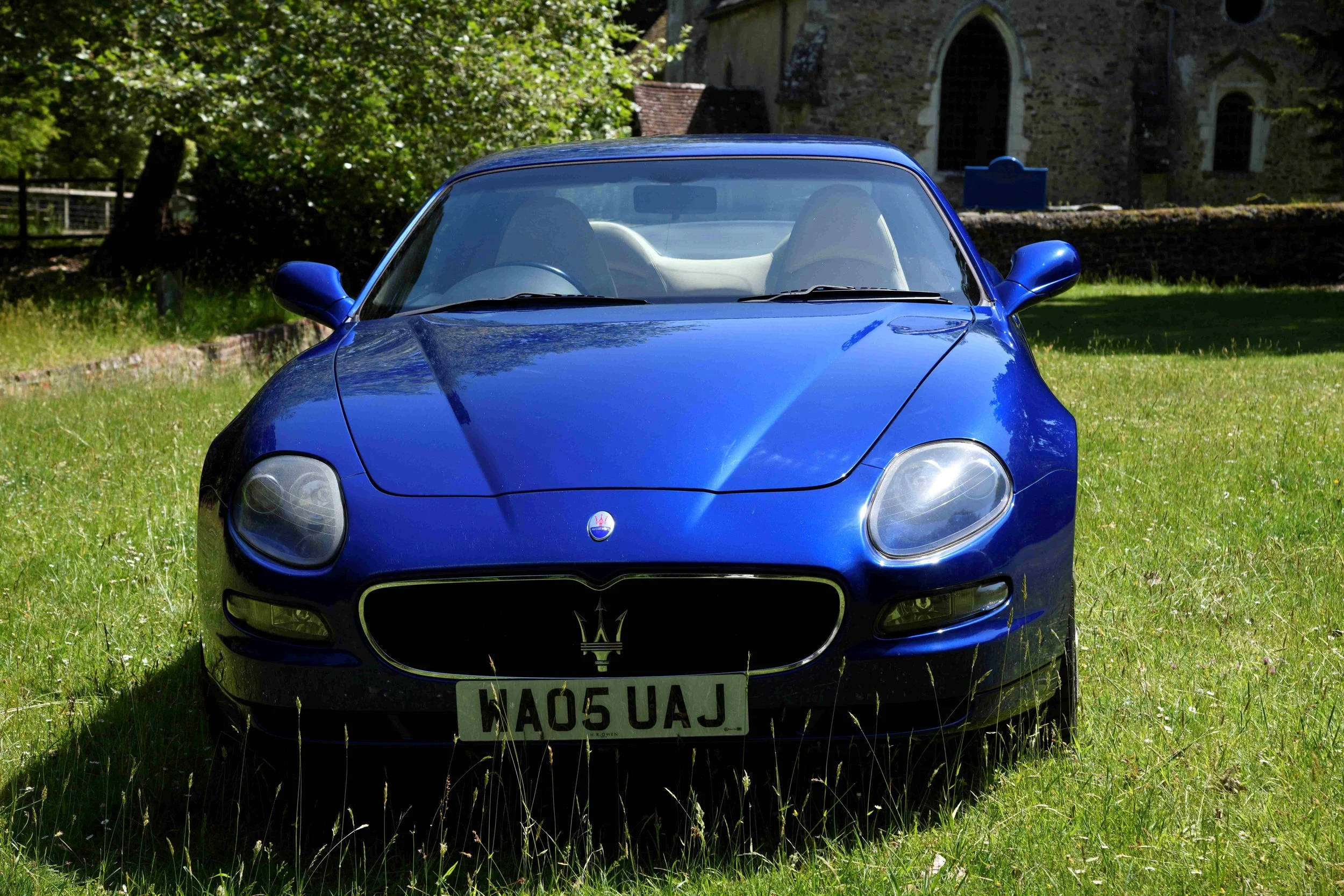 Front view of a blue Maserati convertible car parked on grass with a stone building and trees in the background.